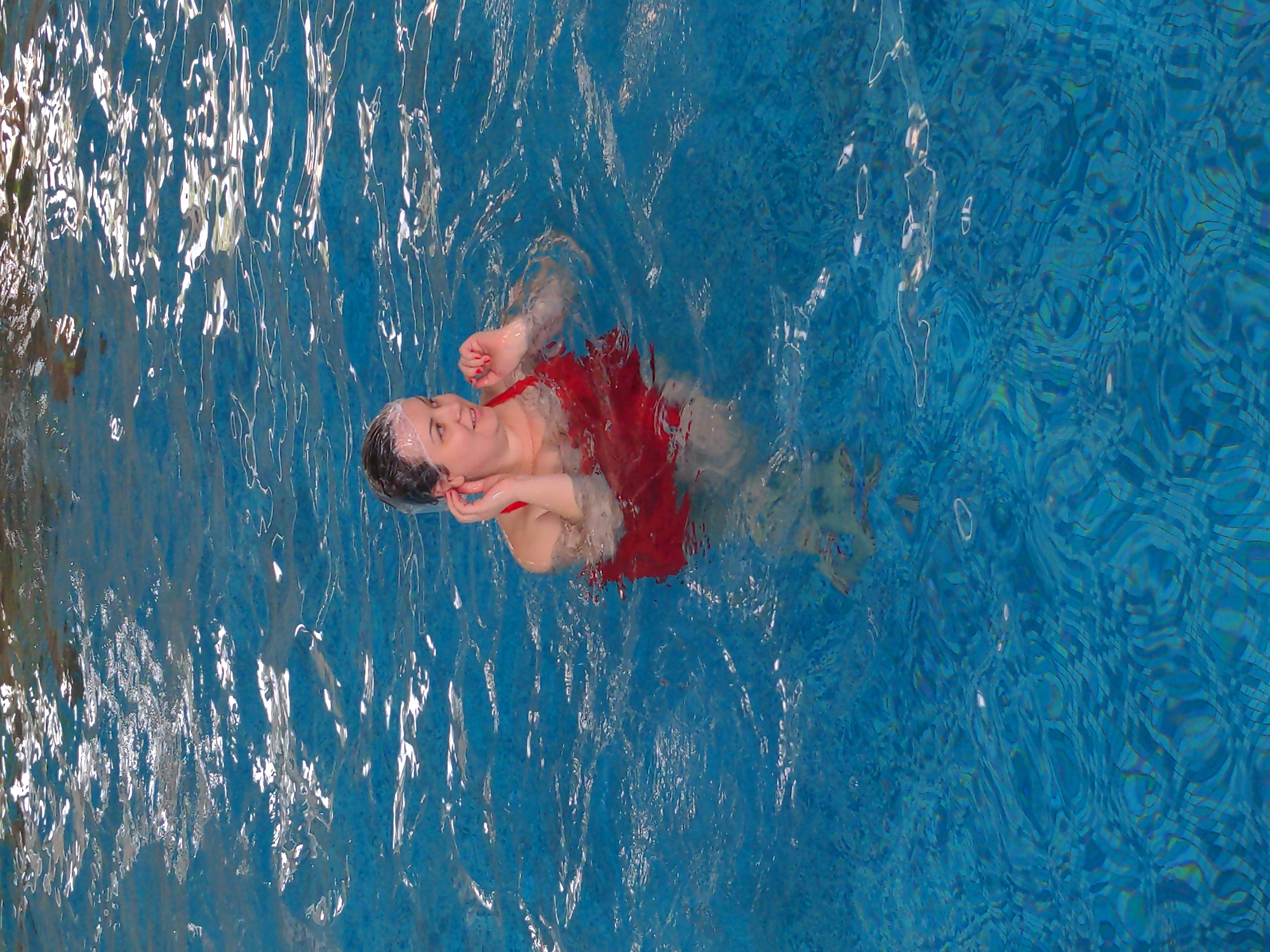 A woman is in a swimming pool in Holubyne, Ukraine. She is wearing a red swimsuit and a swimming cap. The water is blue and there are ripples on the surface. The woman is looking to the right and appears to be smiling. Her hands are near her face. The bottom of the pool is visible and appears to be tiled.