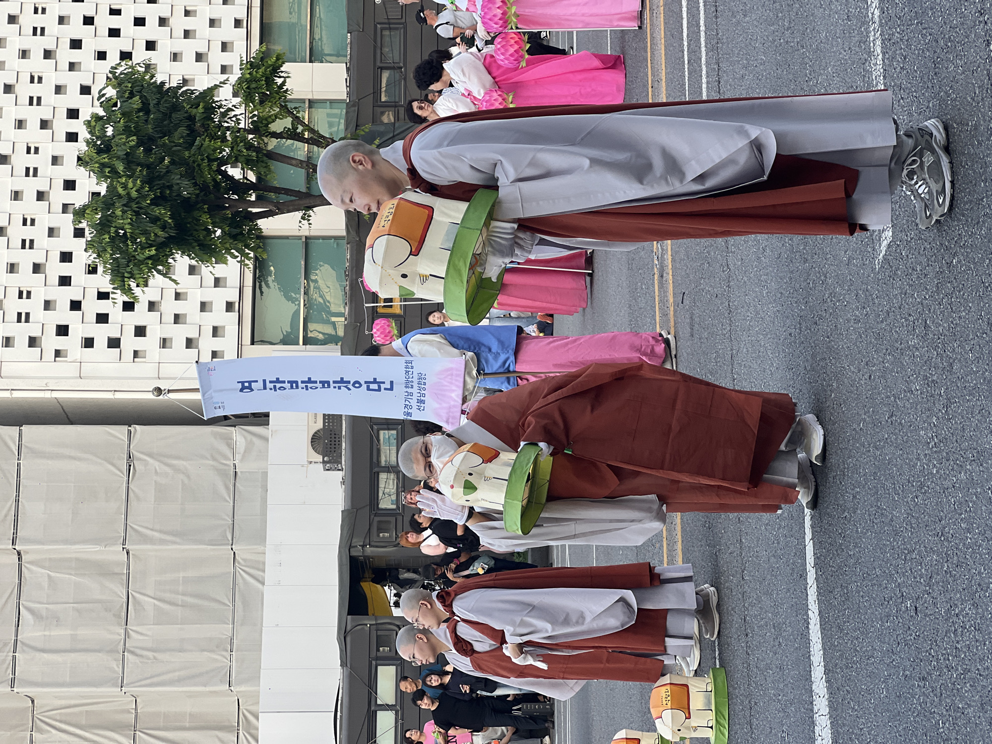 The image shows a row of Buddhist nuns walking in what appears to be a parade or procession in Seoul, South Korea. They wear traditional grey and brown robes and carry lanterns decorated with cartoon images. Some nuns have shaved heads while others wear masks. The street is paved with grey asphalt and has painted white lines. Other individuals, dressed in more casual clothing, can be seen in the background. Buildings are visible, including one with a distinctive grid-like facade. A banner with Korean text is held aloft. A tree is planted near the building. An air conditioning unit of brand "KLASSI" is visible on the building facade.