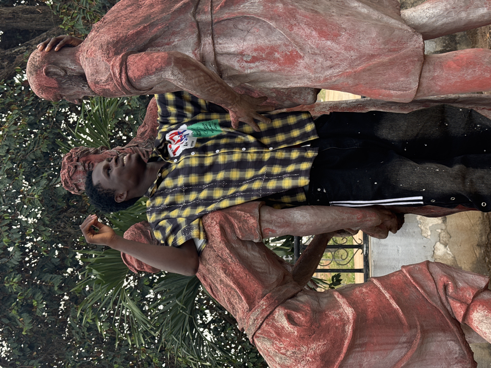 An outdoor scene in Ihiagwa, Nigeria, depicts a young male posing with three weathered, red-painted statues. The male, with short, dark, curly hair, wears a black and yellow plaid button-up shirt featuring a prominent "USA" patch and a green patch with "CHANTING ANTHEMS" text, along with dark pants showing white paint splatters and a vertical white stripe on the right leg. He smiles, looking upwards and to his right. His left arm rests on the back of a crouching statue positioned to his left, while his right side is adjacent to another statue. A third statue, identifiable by its bald head and beard, is positioned to the right, with its right arm extended, its hand resting on the head of the central statue. The statues are made of a rough, grey material partially covered in faded red paint. The background features dense green foliage from trees and palm fronds, while a decorative black metal fence and a concrete base are visible at the lower portion of the image.