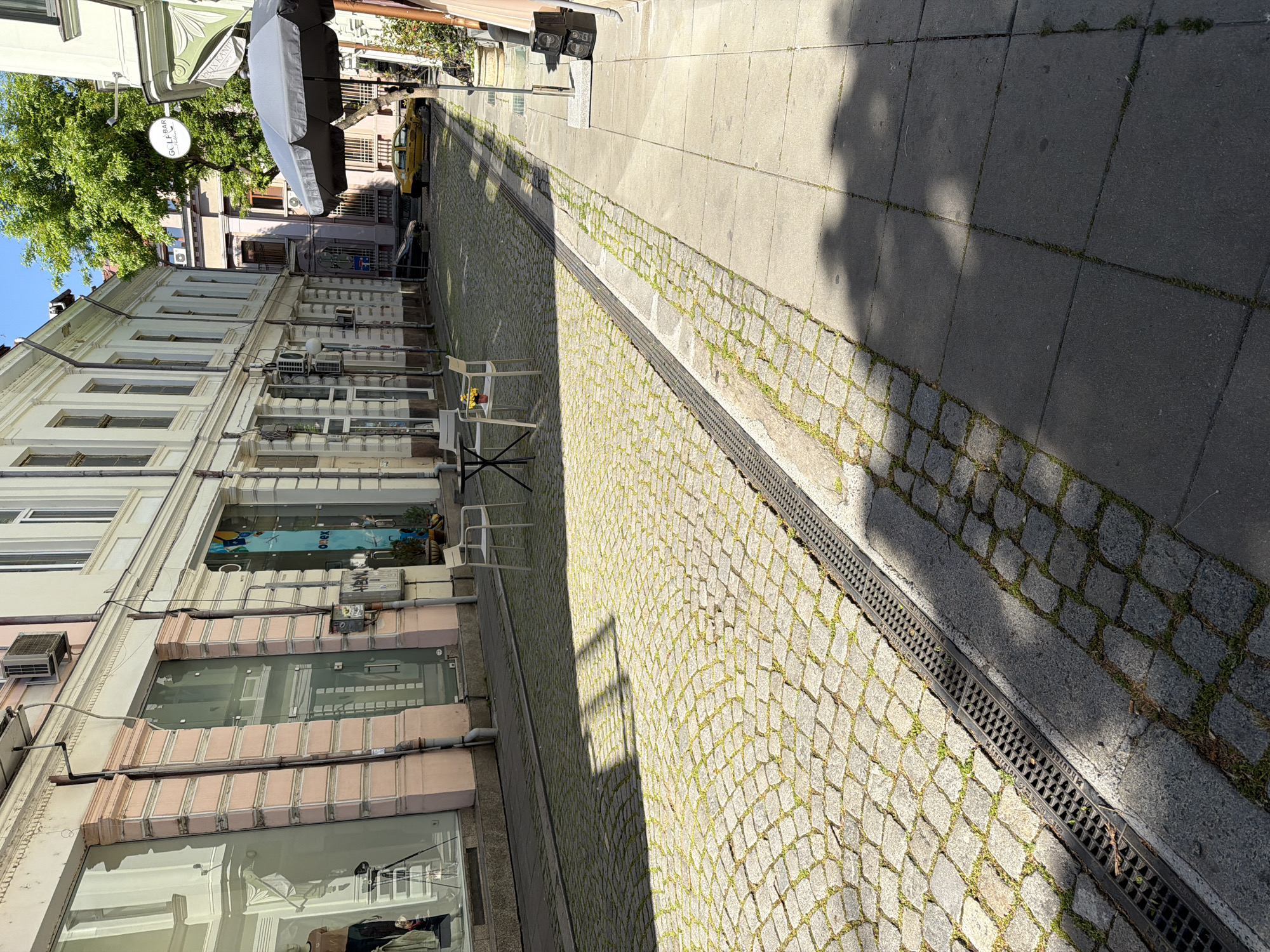 The image depicts a narrow street in Plovdiv, Bulgaria. The street is paved with cobblestones on one side and smooth paving stones on the other, separated by a drainage grate. On the left, a multi-story building with a mix of residential and commercial spaces lines the street. A cafe, denoted by tables and chairs, occupies the sidewalk in front of one of the businesses. In the distance, a parked yellow vehicle is visible. The overall environment suggests a mixed-use urban area with a combination of pedestrian and vehicular traffic. The weather appears to be sunny, with shadows cast on the street.