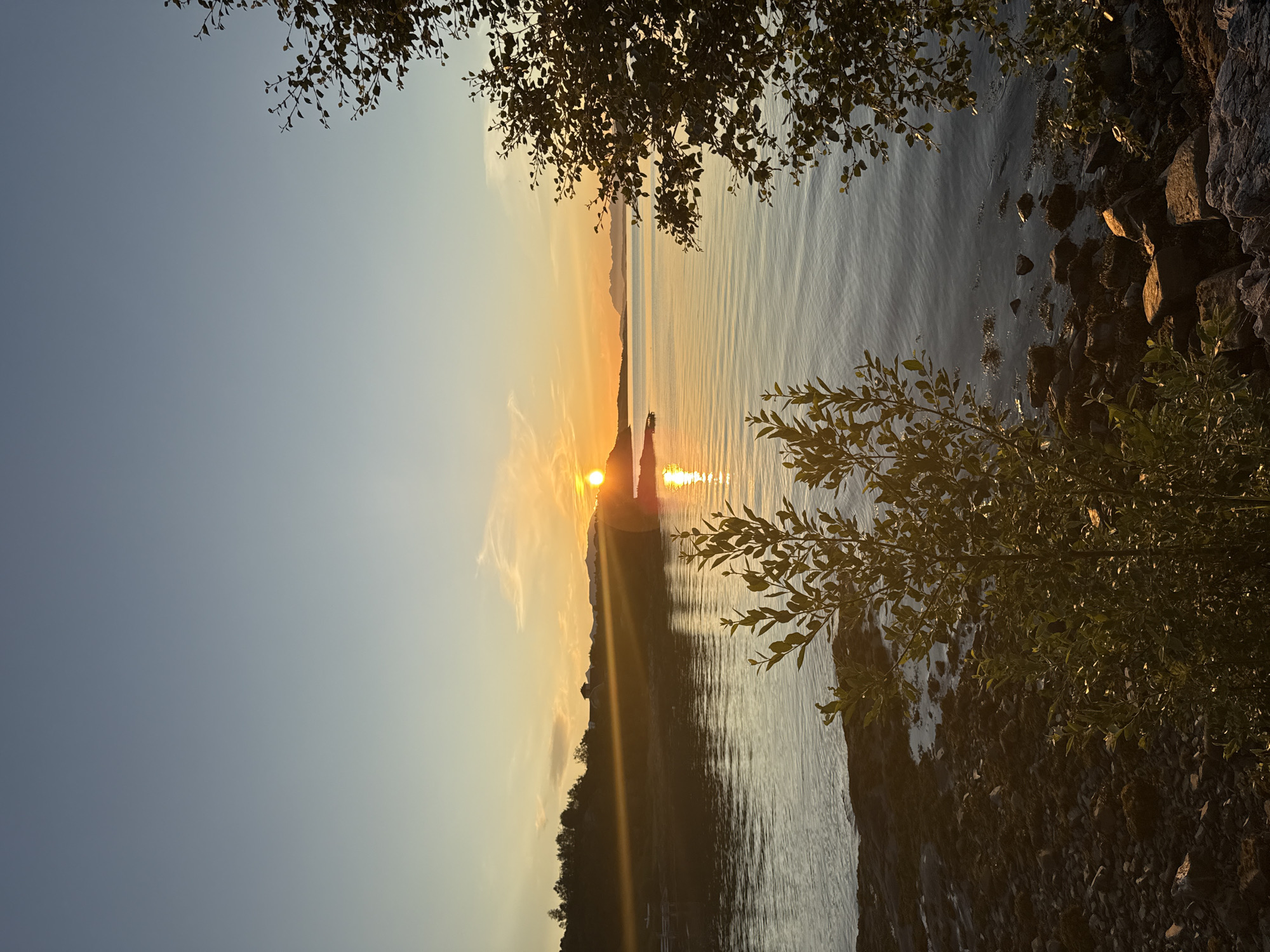 The image depicts a sunset over a calm body of water, likely a fjord or bay, near Harstad, Norway. The sun, a bright orange orb, is positioned low on the horizon, casting a golden glow across the water and illuminating the sky with warm hues of orange, yellow, and pale blue. The water's surface is gently rippling, reflecting the colors of the sunset.
In the foreground, on the right side of the frame, a tree with dark green leaves partially obstructs the view, its branches reaching inwards. On the left side, another tree, also with green foliage, is visible in the lower part of the image, with a rocky shoreline in front of it. The shoreline is composed of various-sized stones and some seaweed.
In the middle ground, a distant, hilly landmass with trees lines the horizon. A few structures, possibly houses or cabins, are visible on this landmass. The sun's rays create a distinct beam of light stretching across the water towards the left side of the image. The sky above is clear, transitioning from a pale blue at the top to warmer tones near the horizon. There are no people or animals visible in the scene. The overall atmosphere is serene and tranquil, characteristic of a northern sunset.