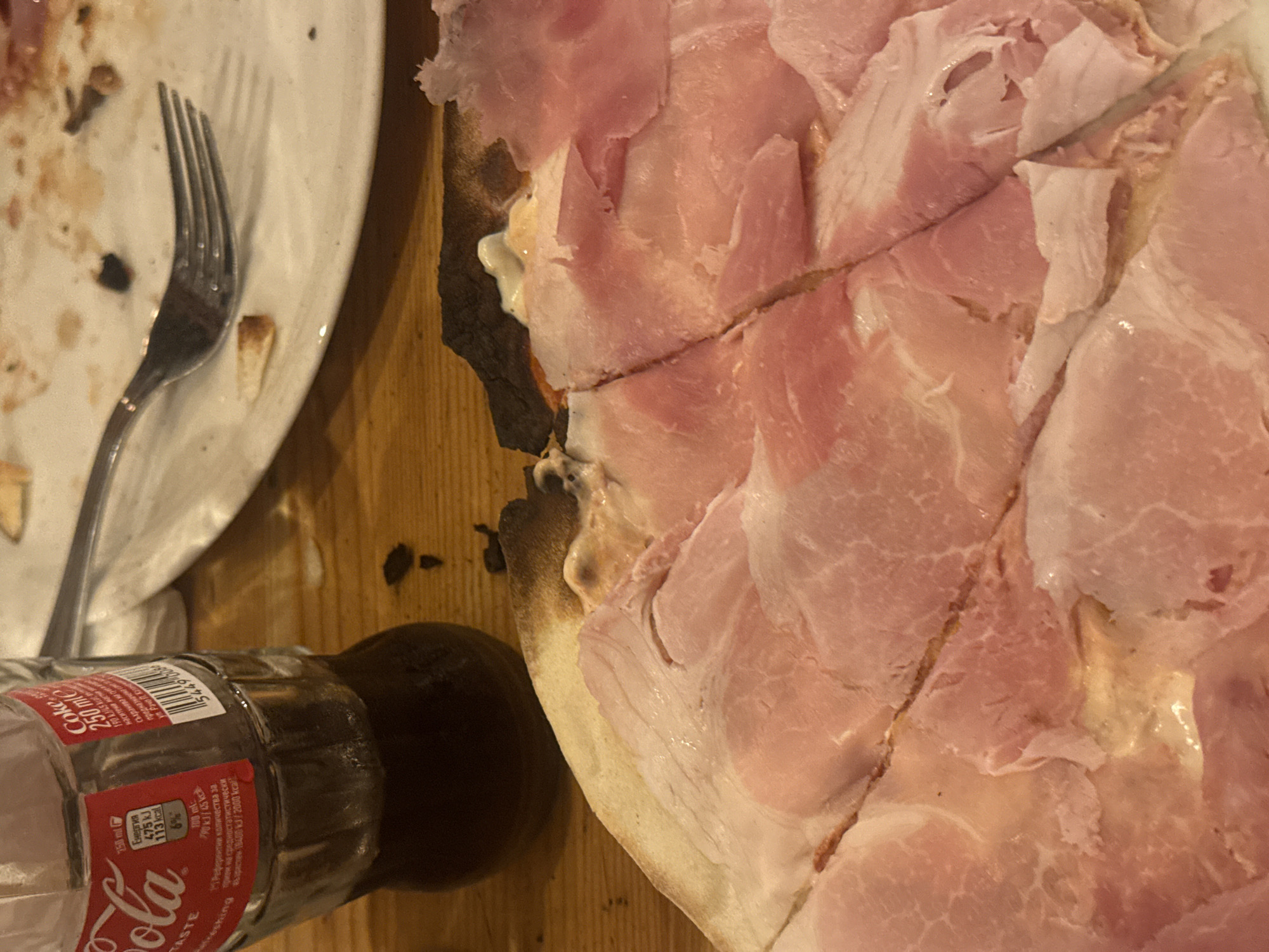The image shows a partially consumed pizza topped with a generous amount of prosciutto. Slices have been cut, and some prosciutto appears to be overlapping. A dark, possibly burnt crust is visible along one edge. Next to the pizza is a partially empty, small, glass Coca-Cola bottle labeled "250 ml". An off-white, used plate with a silver fork sits near the pizza, with food remnants visible on the plate. The entire scene is set on a wooden table or surface. The Coca-Cola bottle has labeling in Cyrillic script, suggesting the location could be in Bulgaria.