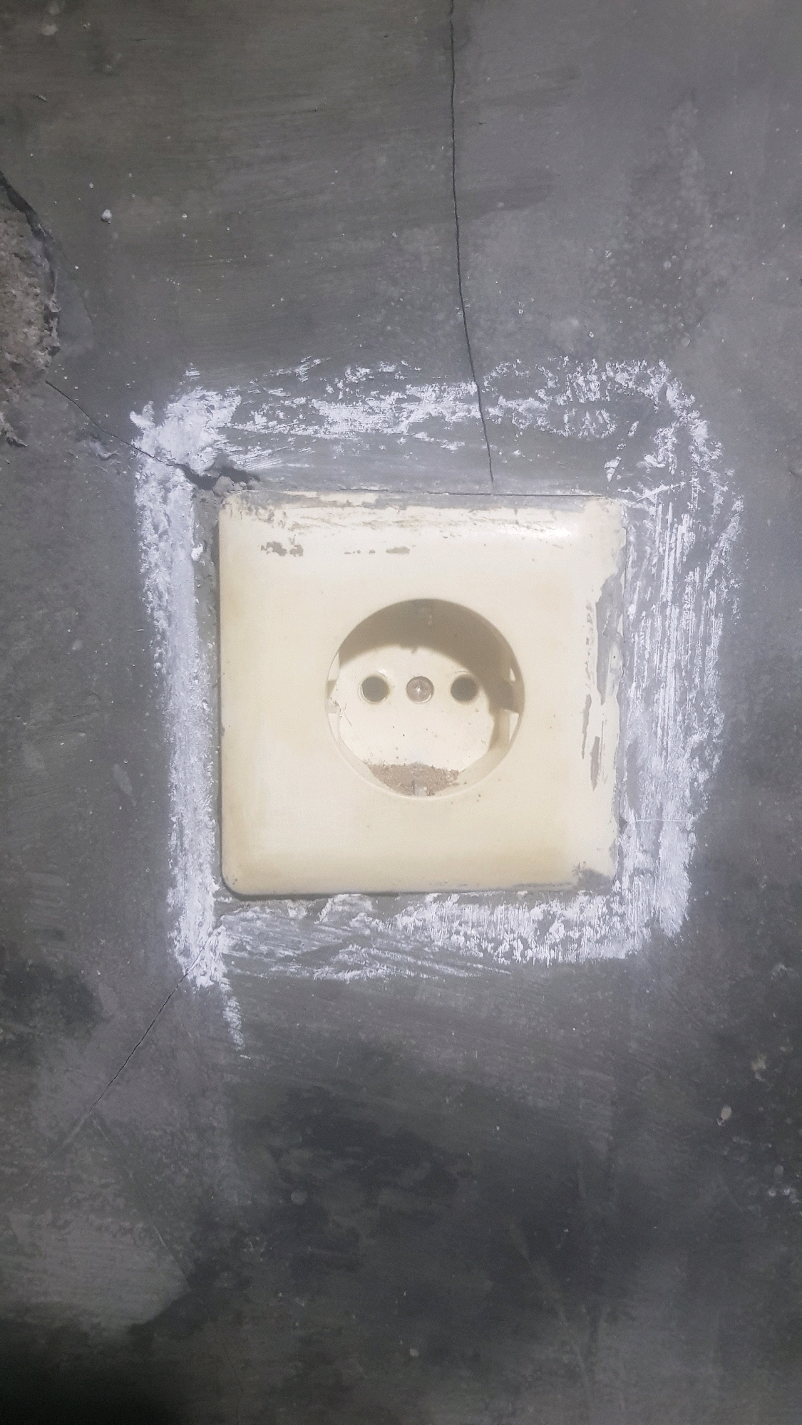 A cream-colored electrical outlet with three circular openings is embedded in a dark gray, cracked concrete wall. The outlet is surrounded by a rough, white substance, possibly plaster or sealant, which is unevenly applied. There are visible cracks in the concrete wall, both around the outlet and extending upwards and to the left. The surface of the wall appears dusty and worn. The outlet itself is slightly discolored and has some debris inside the openings.
