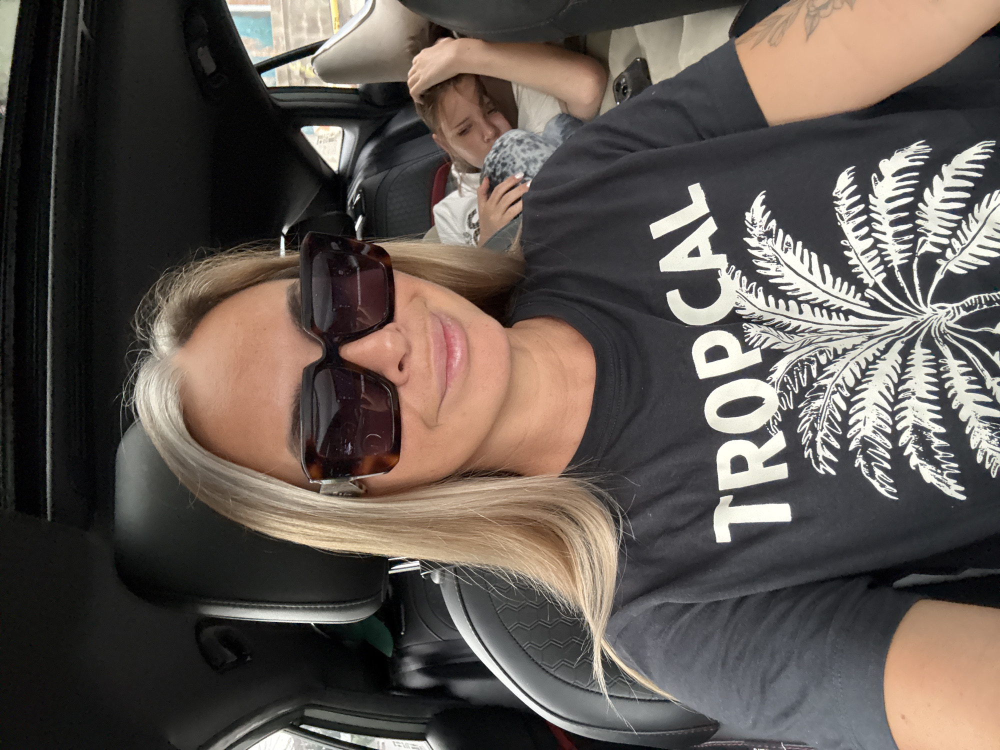 In the city of Ko Samui, Thailand, a woman with blonde hair, wearing large, square sunglasses and a black t-shirt with a white "Tropical" palm tree graphic, is taking a selfie from the front passenger seat of a car. Behind her, in the back seat, a young child is sleeping, with their head resting on a pillow. The car interior appears to be black leather with a sunroof. The lighting suggests it is daytime, possibly overcast.