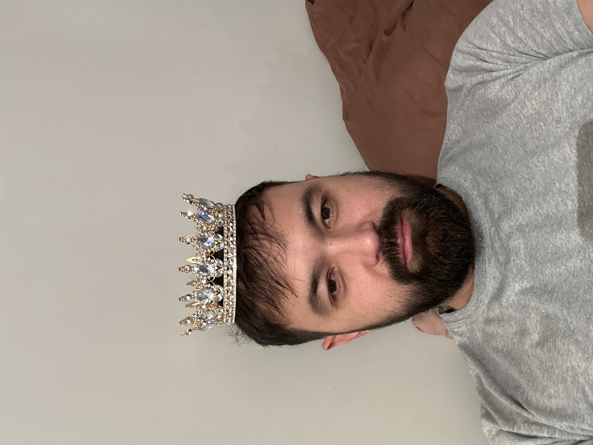 A person with a beard and wearing a gray t-shirt is lying on their side. They are also wearing a jeweled crown. The background includes a white wall and what appears to be a brown blanket or pillow. The location in the city of Khimki, Russia, cannot be determined from the image.