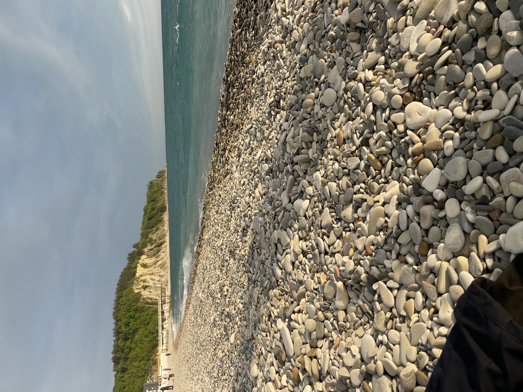The image shows a pebble beach in Olginka, Russia. The beach is covered in smooth, grey and white pebbles. To the left of the pebbles is the Black Sea with its green, foamy waves. Further to the left is a cliff covered in green trees. In the lower left corner of the image, there are several buildings including a pergola or gazebo-like structure. In the bottom right corner of the image is a dark, possibly black, part of a jacket.