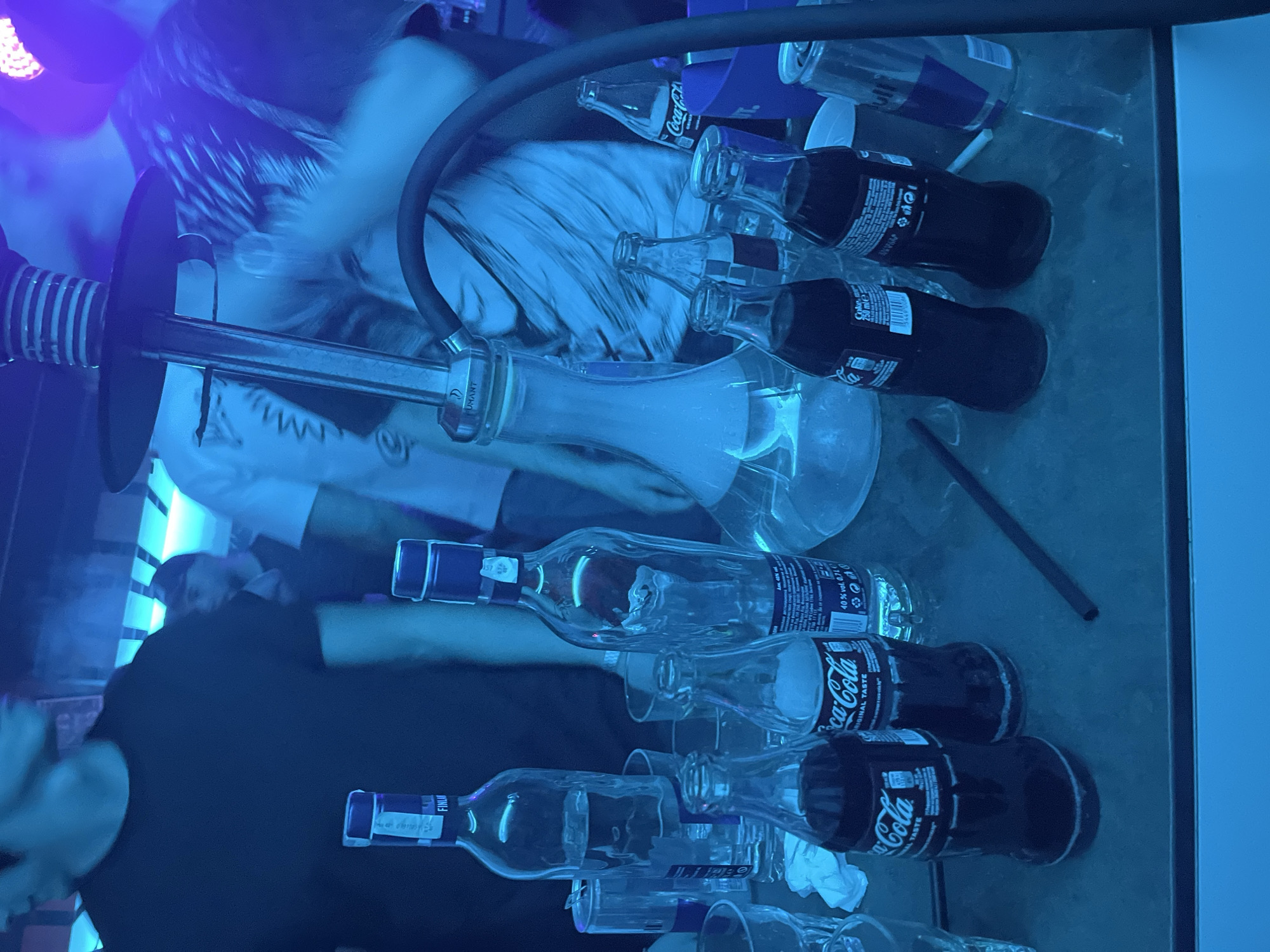 The image depicts a nightlife setting, potentially a bar or club, with a hookah prominently featured. Several individuals are visible, one wearing a white shirt and another in a black shirt. Various drink containers, including bottles of Coca-Cola, Red Bull cans, and empty liquor bottles with labels "FINLANDIA" are strewn across a reflective surface, suggestive of a table or bar top. Empty glasses are scattered among the bottles. The overall lighting in the scene is blue, creating a club-like atmosphere. The location and city cannot be determined.