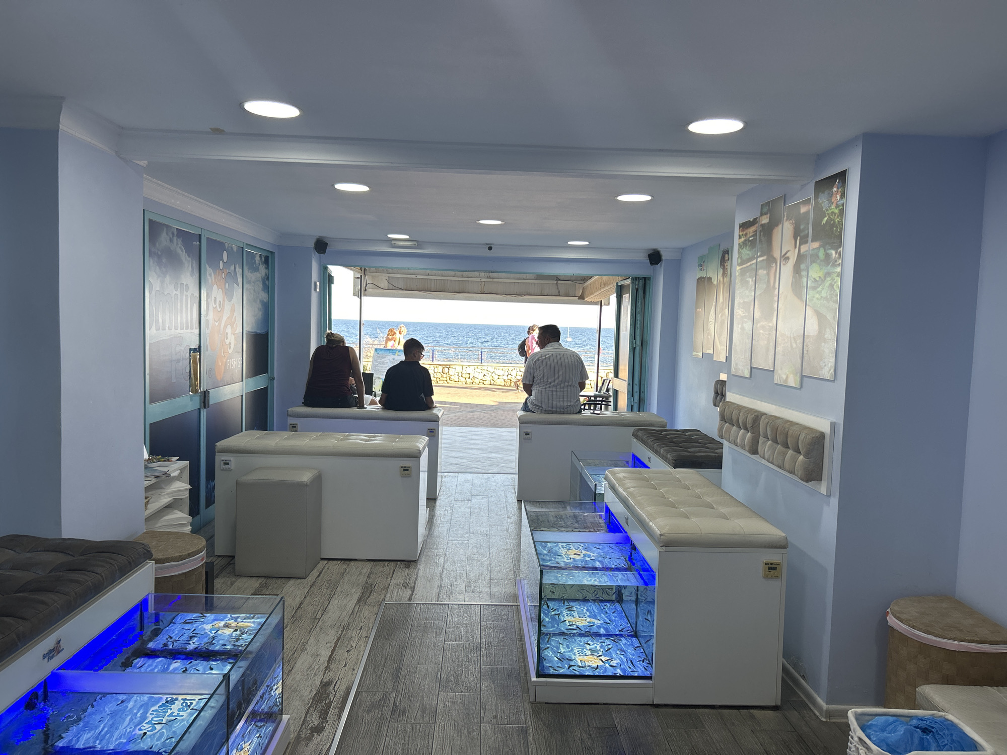 The image depicts the interior of a fish spa located in Cala Rajada, Spain, with a view of the Mediterranean Sea visible through an open doorway at the far end. The room is painted a light blue and features rows of white benches with cushioned seating above clear glass tanks filled with water and small fish, commonly used for fish pedicures. Several individuals are seated on the benches, with their feet submerged in the tanks.
In the foreground, a dark grey cushioned bench is positioned above a glass tank illuminated with blue light, containing numerous small fish. To the right, a white bench sits atop a similar fish tank, also illuminated. Further back, more white benches and fish tanks are visible, some with people participating in the fish spa treatments.
Through the open doorway, the blue expanse of the sea and a stone-paved walkway are visible, suggesting a beachfront location. The flooring inside the spa consists of grey wooden planks, and the walls are adorned with framed artwork or decorative panels. A white storage unit with compartments is visible on the left side of the room. The overall atmosphere suggests a relaxing and therapeutic environment.