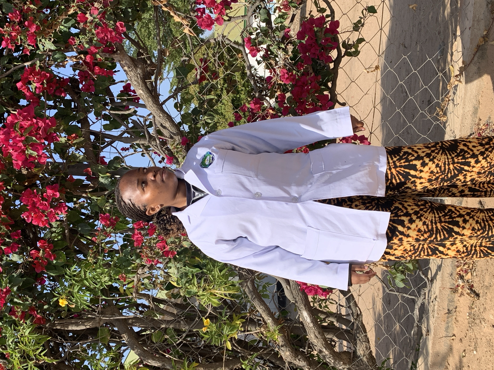 A young Black woman stands in front of a blooming bougainvillea tree in Dodoma, Tanzania. She is wearing a white lab coat over a dark shirt, with black and orange patterned pants. A wire mesh fence is visible in the foreground, and a building with a green roof can be seen in the background. The woman is looking to her right, and her hair is styled in braids. The lighting suggests it is a sunny day.