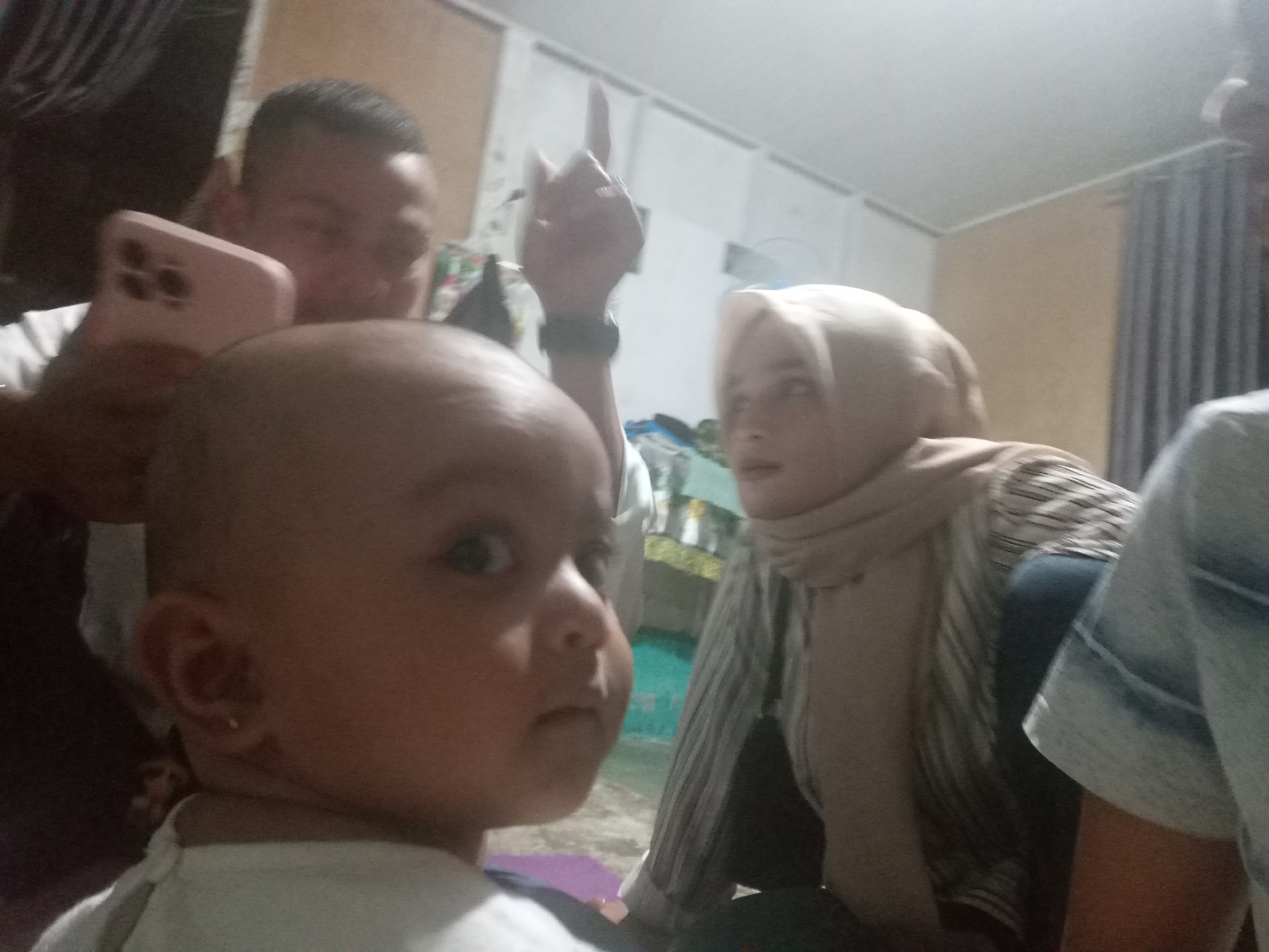 The image captures a close-up of a baby's face in the foreground, looking directly at the camera with wide eyes and a slightly open mouth. The baby is wearing a white garment. Behind the baby, to the left, an adult's head and torso are partially visible, holding a light pink smartphone. The adult is looking down at the phone. To the right of the baby, another adult is visible, wearing a headscarf and a striped shirt. This adult is looking towards the camera. A third person's arm, wearing a denim jacket, is also visible on the far right of the frame. The background appears to be an indoor setting with plain walls and curtains. The lighting is dim, suggesting it might be evening or indoors with artificial light. The overall scene depicts a casual, possibly family gathering.