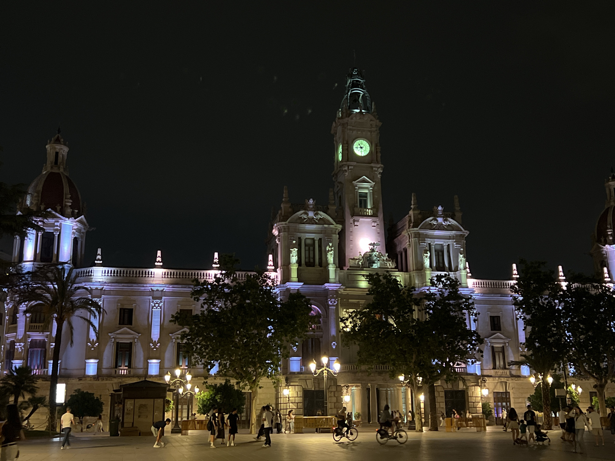 The image depicts a nighttime scene in the Plaça de l'Ajuntament (City Hall Square) in València, Spain. The València City Hall is the prominent architectural feature, illuminated with purple, blue, and white lights, its clock tower showing a green-lit clock face. Trees line the square, partially obscuring the lower sections of the building. Streetlights provide additional illumination. Pedestrians are visible throughout the square, including individuals walking, standing, and riding bicycles. The ground surface appears to be paved and reflective.