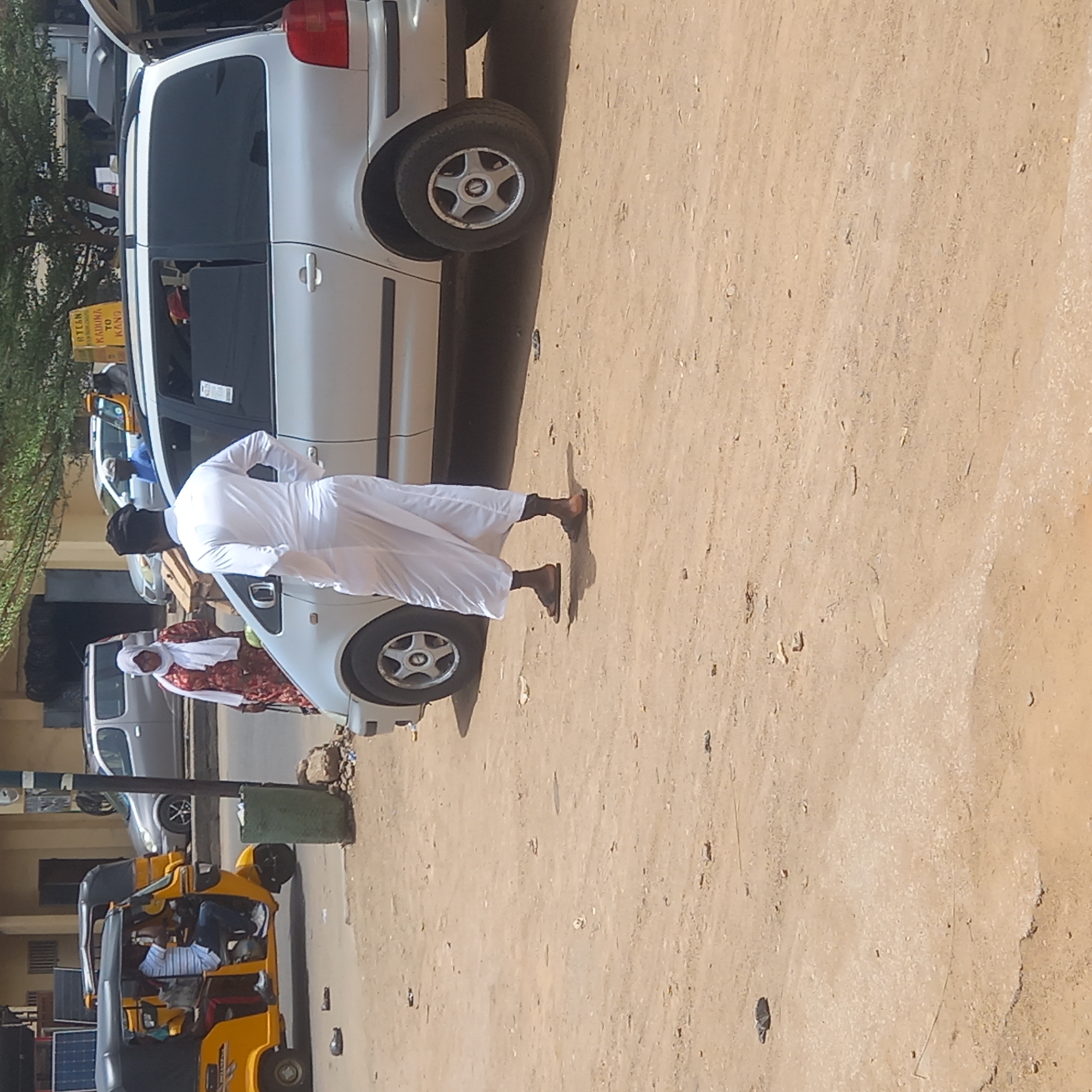 The image captures an outdoor street scene in Dadi Riba, Nigeria, characterized by bright daylight and a dusty, light brown ground. In the mid-ground, a man dressed in a long white garment, a black cap, and sandals stands with his back to the viewer, facing a parked white hatchback car. The car's rear trunk is open, and a black suitcase or bag is visible inside. Another person, possibly a woman, wearing a patterned dress and a headscarf, is partially visible behind the white car, near its front wheel.
Further to the left, a yellow auto-rickshaw, commonly known as a Keke Napep, is parked. A person wearing a striped shirt and jeans is seated in the driver's position. Near the auto-rickshaw, what appear to be solar panels are visible. In the background, other vehicles, including additional white cars, are parked, and green foliage from trees can be seen on the left.
A notable detail is a yellow sign with black text visible on the left side. The text clearly reads "RTEAN" at the top, followed by "KADUNA TO KANO" below it. This indicates the location is likely a motor park or a public transport hub, with services to the Nigerian cities of Kaduna and Kano, possibly operated by the Road Transport Employers' Association of Nigeria (RTEAN). The scene suggests a typical day at a local transport point.