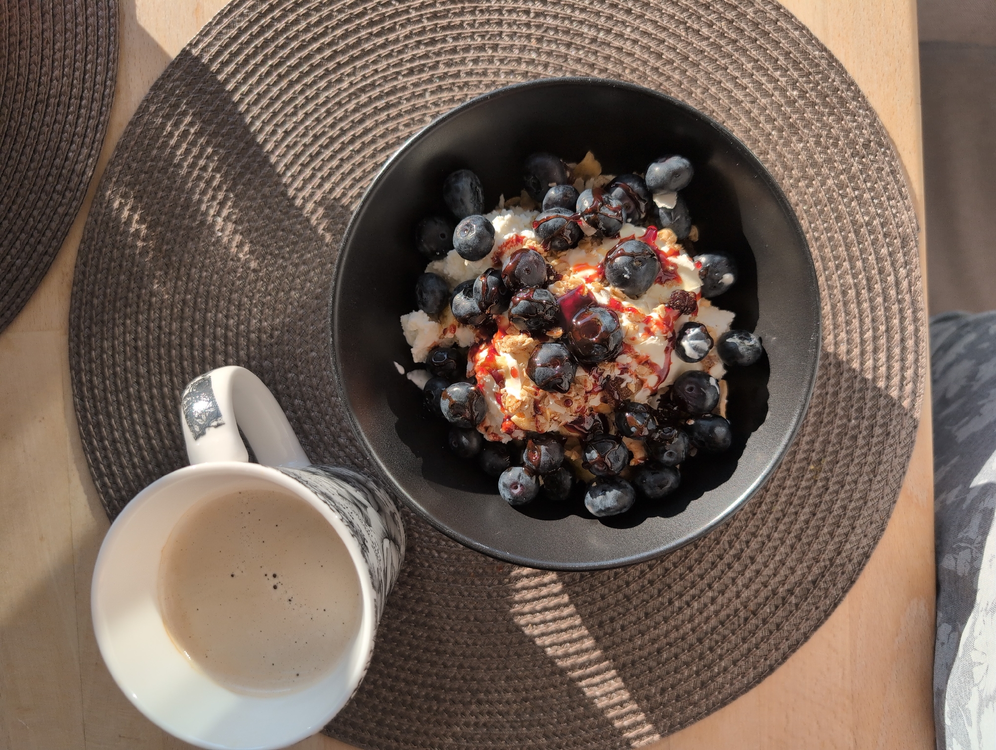 A overhead, high-angle medium shot shows a breakfast meal and a cup of coffee, positioned on a textured, circular placemat on a wooden surface. The placemat is dark brown, with a woven texture. To the left, a white coffee mug with a black graphic pattern is filled with light brown coffee, topped with a thin layer of foam. To the right of the mug, a black bowl contains a white creamy substance, possibly yogurt or cottage cheese, topped generously with fresh blueberries. Drizzled over the blueberries and creamy substance is a red syrup and some crushed nuts or granola. Sunlight casts diagonal shadows across the placemat and the wooden surface. The background is blurred, but appears to be a wooden table. There are no people visible in the image. The scene depicts a simple breakfast setting. The location is not specified in the image or its metadata.