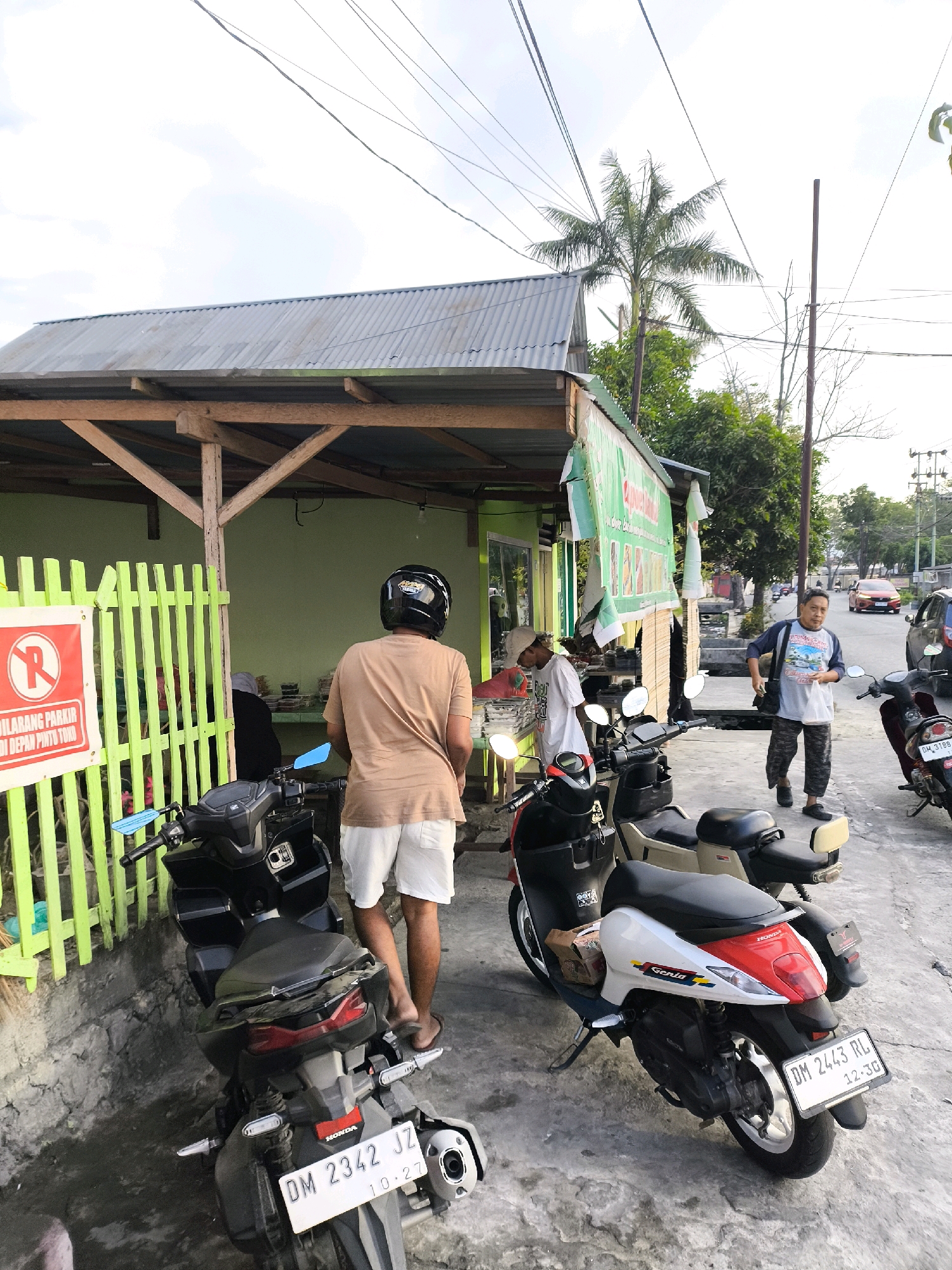 This is a street-level, eye-level, medium shot taken in Gorontalo, Indonesia. In the foreground, there are two motorcycles parked side-by-side on a concrete pavement. The motorcycle on the left is black, and the one on the right is white and red. Behind the motorcycles, a man wearing a brown t-shirt and white shorts, with a black helmet on, stands facing away from the camera. To the left of the motorcycles, a bright green wooden fence is visible, with a red and white sign attached to it displaying a red circle with a diagonal line through a red "R," indicating no parking. Further back, a man in a light-colored t-shirt and patterned pants walks away from the camera. He is carrying a bag over his shoulder. Adjacent to the fence is a building with a green wall and a corrugated metal roof supported by wooden beams. The building appears to be a shop or food stall, with items displayed on shelves and tables. A palm tree and other vegetation are visible in the background, along with power lines and a street with cars. The lighting suggests it is either early morning or late afternoon.