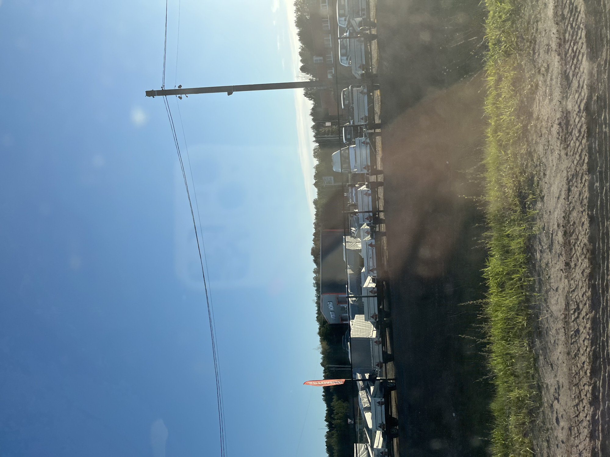 The image shows a horizontal view of an area near Chaikovsky, Russia, taken during the day. On the right side is a dirt road with grass on its edge. Adjacent to the road are multiple parked trailers. Across from the trailers are trees. Overhead, a blue sky is visible with electrical wires. The location includes visible buildings of Chaikovsky, Russia.