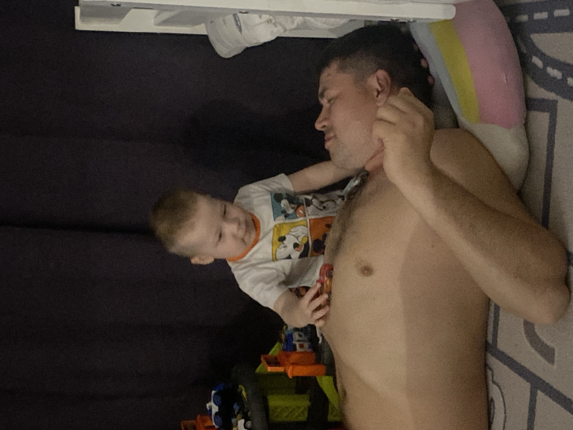 A fair-skinned adult male lies shirtless on a bed with a multi-colored striped pillow to his right, possibly in a private residence in Sochi, Russia. The male's right hand is raised to his neck. A young child wearing a long-sleeved shirt with cartoon characters is leaning over the man. Toys are scattered on the bed near the child, including orange and green objects. A dark curtain is visible in the background. The bed has a gray and white patterned bedsheet. A shelf is partially visible above them.