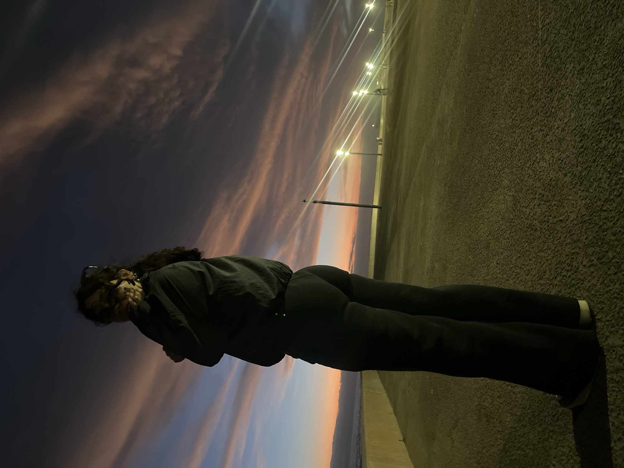 A full-body shot of a woman in a black jacket and pants is captured against a dramatic sky. She is standing on a concrete surface, looking back at the camera. The location appears to be along a coastal road or promenade in Arica, Chile, evidenced by the presence of a string of street lights lining the road. The sky displays a vivid sunset, transitioning from darker hues to lighter shades of pink and orange closer to the horizon. Part of what appears to be the ocean is visible in the bottom of the frame.