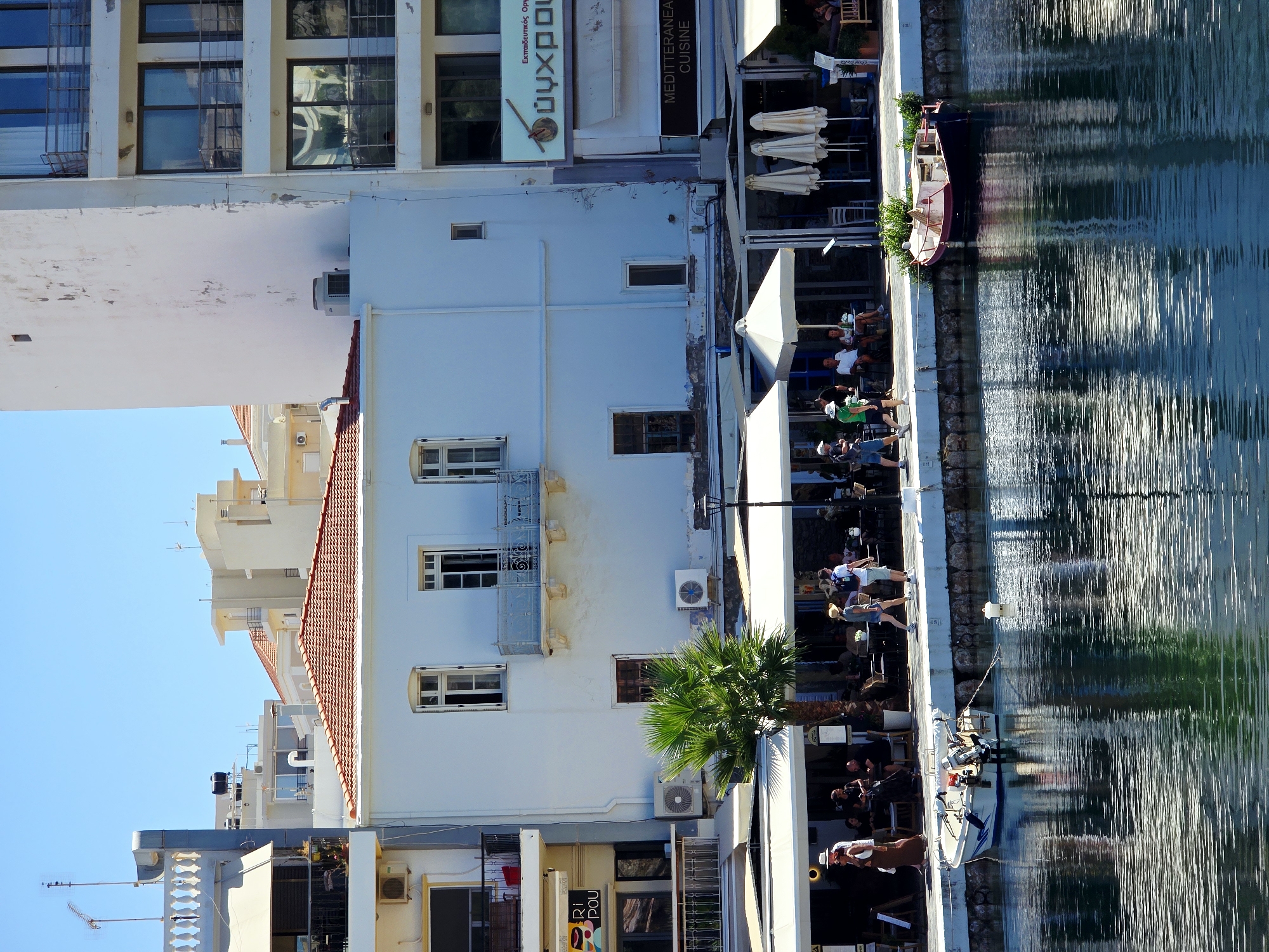 This is a medium shot of a waterfront promenade in Agios Nikolaos Municipal Unit, Greece. The scene is characterized by buildings, people, boats, and water.
In the background, there are several multi-story buildings. The building in the center is painted white with a red tiled roof. It has several windows and balconies. To its left is a lighter colored building with a balcony and an air conditioning unit. To the right of the central building is a taller, white building with dark-rimmed windows and balconies. Signage in Greek is visible on this building.
In the foreground, the water of a harbor is prominent, reflecting the sky and buildings. A small white boat with an outboard motor is tied to a white line floating in the water. Another boat, darker with a red hull, is moored closer to the shore.
Along the promenade, there are several people walking and sitting at tables outside cafes and restaurants. A woman in a white hat and a long brown dress is walking towards the left. A man in a white shirt and shorts with a backpack is walking towards the right. Other people are seated at tables, some under white umbrellas. A palm tree stands near the center of the promenade.
The overall atmosphere is sunny and bright, suggesting a daytime scene on a warm day. The architecture and presence of a harbor indicate a coastal town or city.