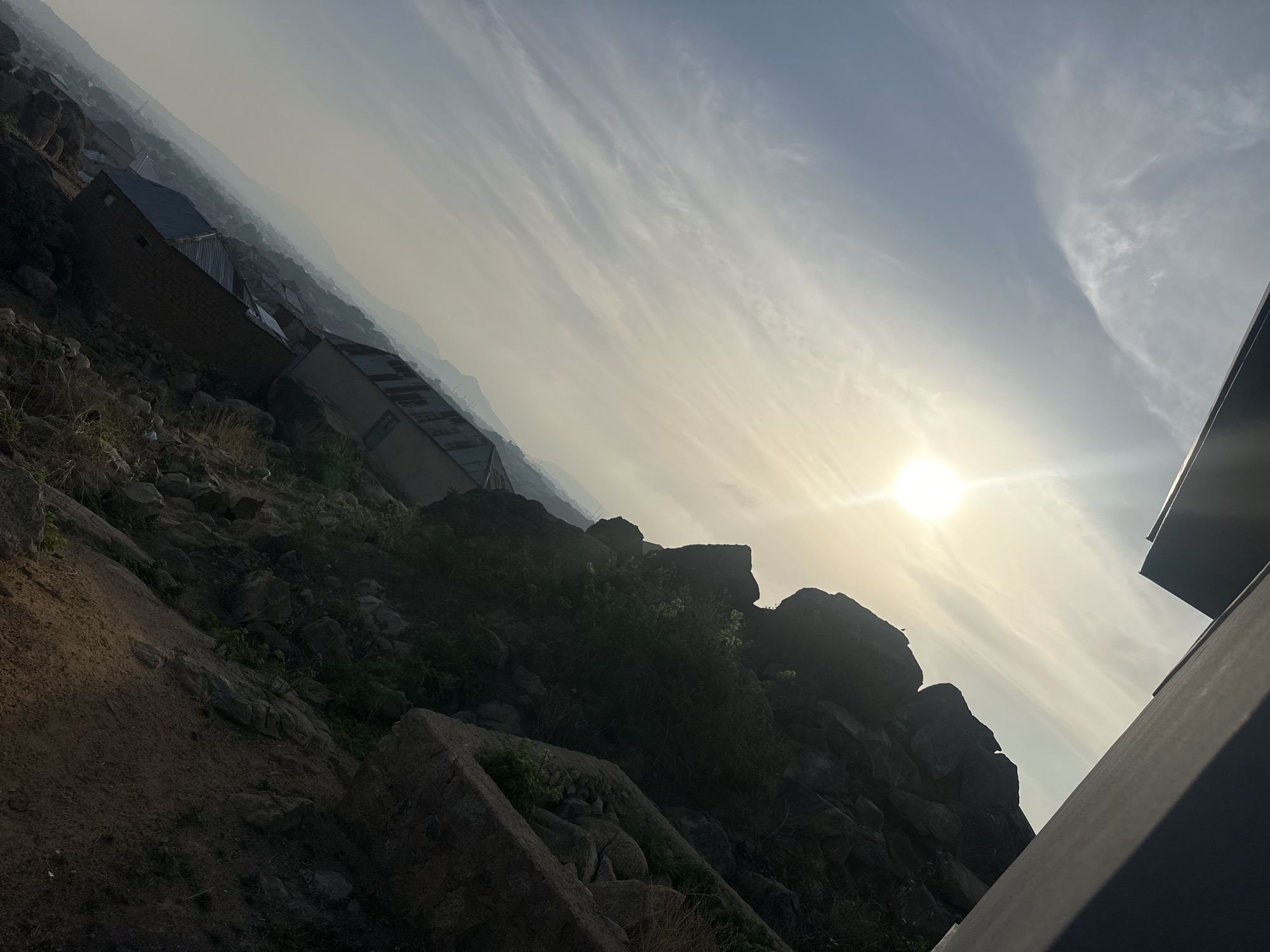 The image presents a tilted view of a landscape in Jos, Nigeria, under a bright sun. The scene includes a mix of natural and man-made elements. The foreground is dominated by a rocky terrain with sparse vegetation, possibly a hillside. Some small structures, including buildings with corrugated metal roofs, are visible on the left side of the frame, suggesting a built-up area in the distance. The sky is bright with sunlight piercing through thin clouds, casting a hazy glow over the scene. A portion of a modern building's structure is visible in the bottom right corner. The overall impression is a blend of urban development amidst a rocky, hilly environment, characteristic of the Jos region.