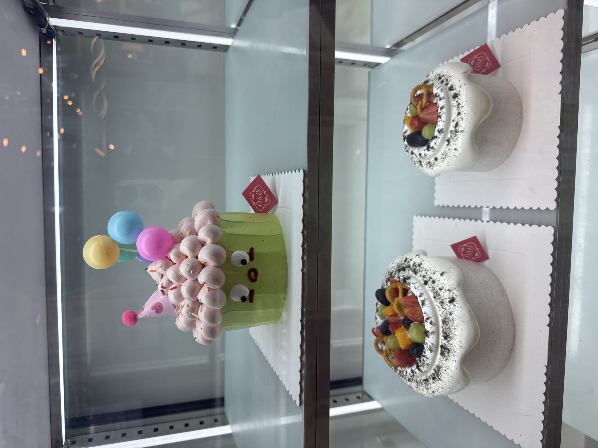 The image shows a display case with two shelves holding cakes. On the left shelf is a green-frosted cake decorated with pink meringue balls, a pink party hat, and balloon decorations. On the right shelf, there are two round cakes with white frosting and black speckles around the edges, topped with fresh fruit. The location appears to be a bakery or cake shop, likely in Đà Nẵng, Vietnam, based on the style of the cakes.