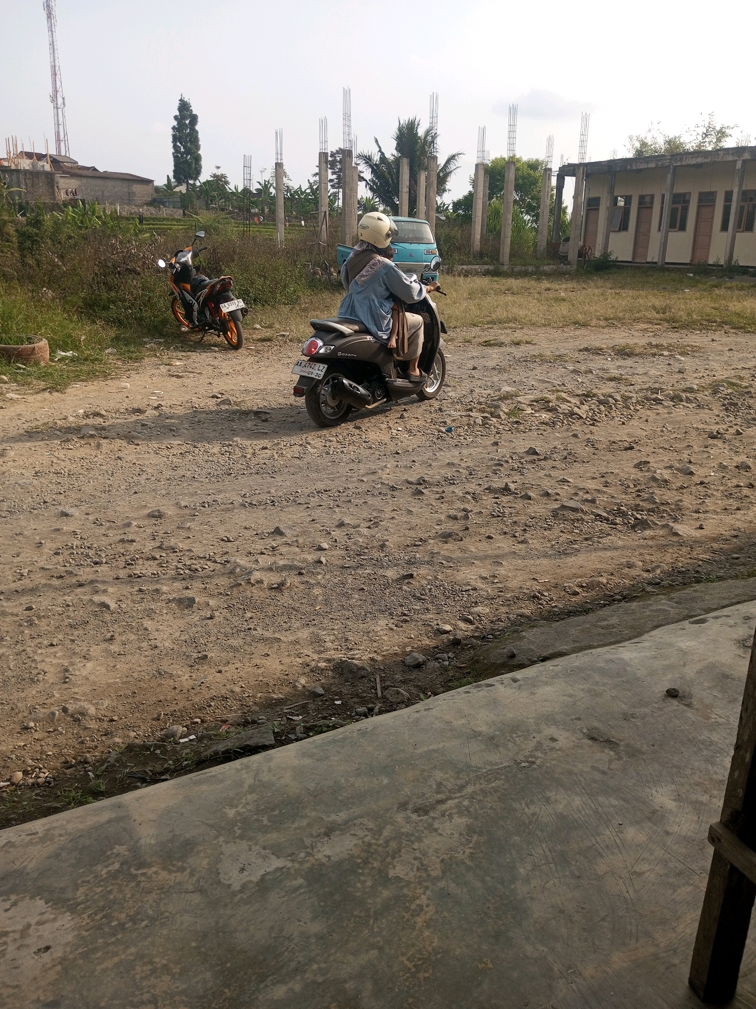 In the city of Kalibeber, Indonesia, a person wearing a light blue top and a beige headscarf is riding a dark-colored scooter on a dirt road. The scooter has a license plate that reads "AA 4742 LZ". Another orange and black motorcycle is parked to the left of the road. In the background, there is a partially constructed building with concrete pillars, a light blue van, and some vegetation. The ground is covered in gravel and dirt. The overall environment appears to be an outdoor, semi-urban area.