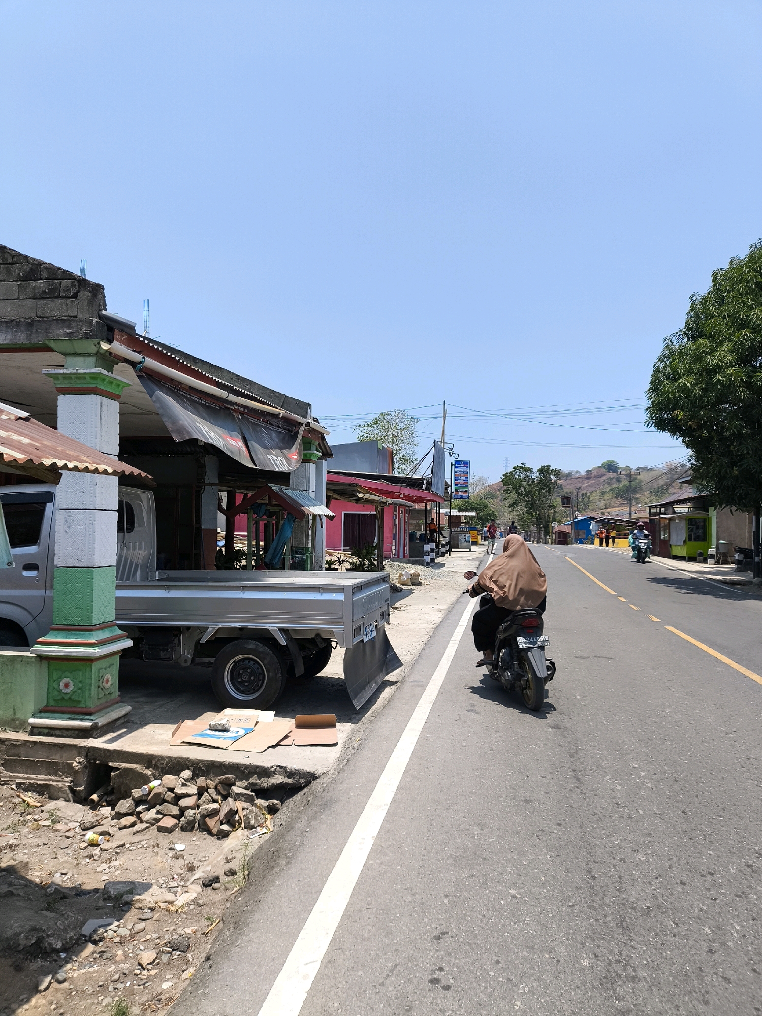 A person wearing a brown hijab rides a motorcycle down a paved road in a rural or suburban area of Indonesia. The road has a yellow center line and a white line on the right edge. The motorcycle is moving away from the camera. On the left side of the road, there is a silver flatbed truck parked next to a building with a partially covered awning. A colorful pillar with green and white decorative elements stands in front of the building. The ground next to the building is unpaved and has piles of rocks and debris. On the right side of the road, there are more buildings, trees, and a hill in the background. The sky is clear and blue, indicating a sunny day.