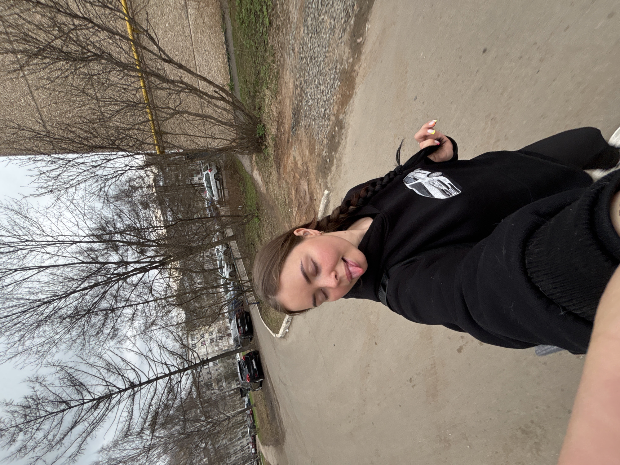 A young woman stands on a paved path, possibly a sidewalk, near a residential area in Izhevsk, Russia. She is wearing a black hoodie with a white car graphic on it. She is sticking her tongue out, suggesting she may be playful. She has a braid.
The environment includes bare trees and parked cars along the street. There is also an apartment building in the background. The weather appears overcast.