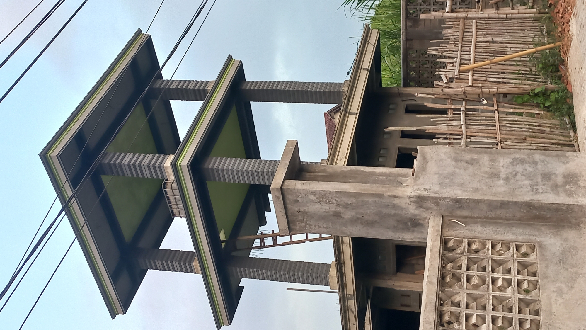 This is a low-angle, vertical photograph taken outdoors of a multi-tiered structure in the city of Pare, Indonesia. The structure appears to be under construction or recently built, with elements of unfinished building visible at the lower levels. The main focus is on the upper sections, which are designed with square pillars and flat, cantilevered roofs. The pillars are decorated with a pattern of alternating dark grey and light grey horizontal bands. The underside of the roofs is painted a lime green color, with dark grey trim along the edges.
The structure is partially obscured by a concrete pillar in the foreground, which is on the left side of the image. To the right of the pillar is a fence made of vertical bamboo poles, some of which are tied together with string. A single bamboo pole leans against the fence. Behind the fence, part of a building is visible, with unfinished walls and windows. Green foliage, possibly sugar cane, is visible behind the fence on the right side of the image. Overhead, dark power lines crisscross the sky, which is overcast with a pale blue-grey hue. A wooden ladder is propped up on the structure, leaning against one of the pillars. A small, cage-like structure is visible on the top level of the building. The ground in the foreground is a rough, paved surface.