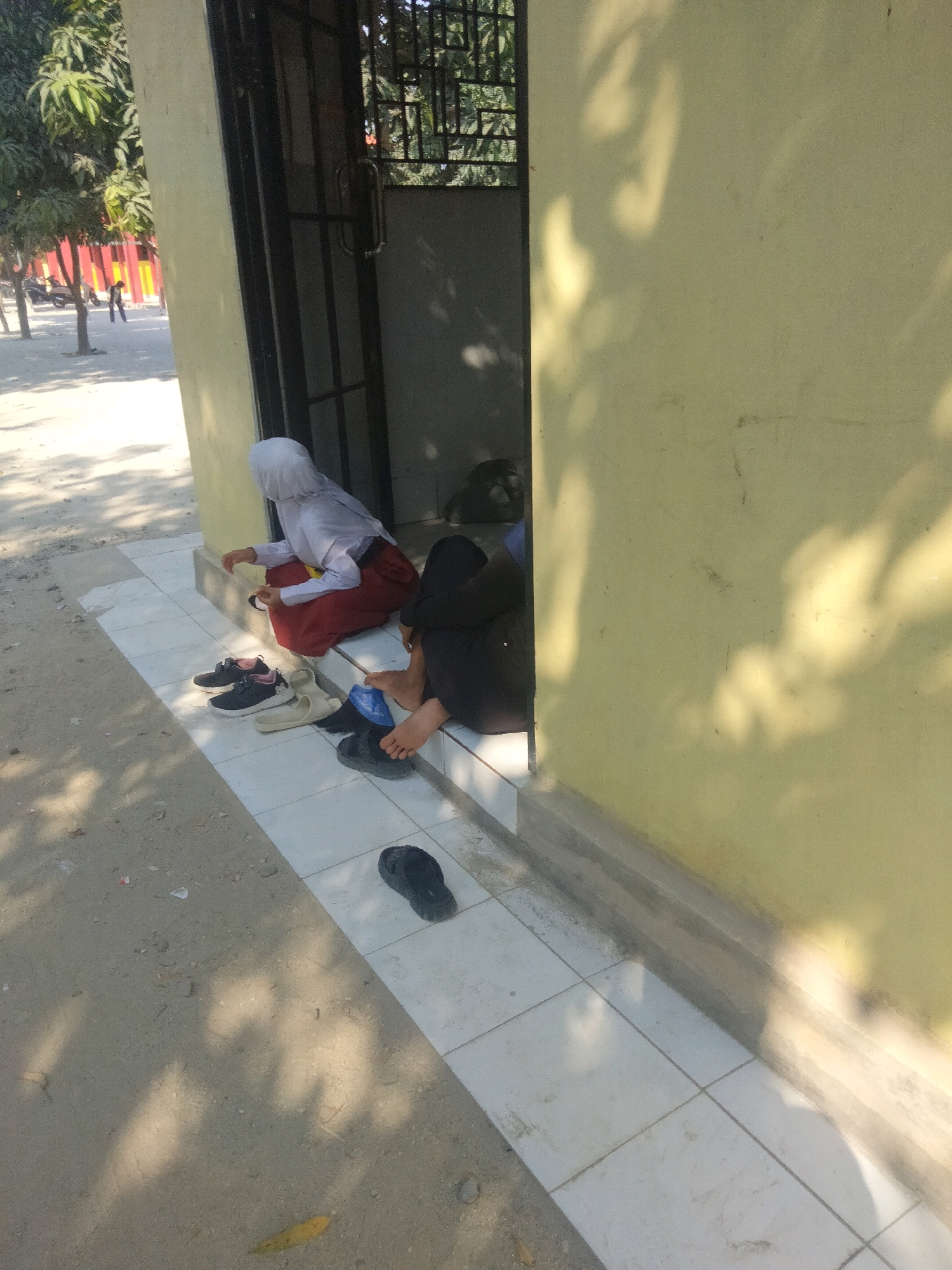 Two individuals are seated outdoors next to a yellow wall, adjacent to a doorway with a metal gate. One person, wearing a white hijab and a red skirt with a white shirt, is facing away from the camera, seated on a tiled surface. Next to them, a second person, wearing dark clothing, is also seated on the tiled surface, facing towards the right of the frame. Several pairs of footwear, including black sneakers and beige sandals, are placed on the tiles in front of the individuals. A black sandal is also visible on the sandy ground to the right. The environment appears to be an outdoor area with trees and shadows cast by sunlight. The location is identified as Suka Sari, Indonesia.