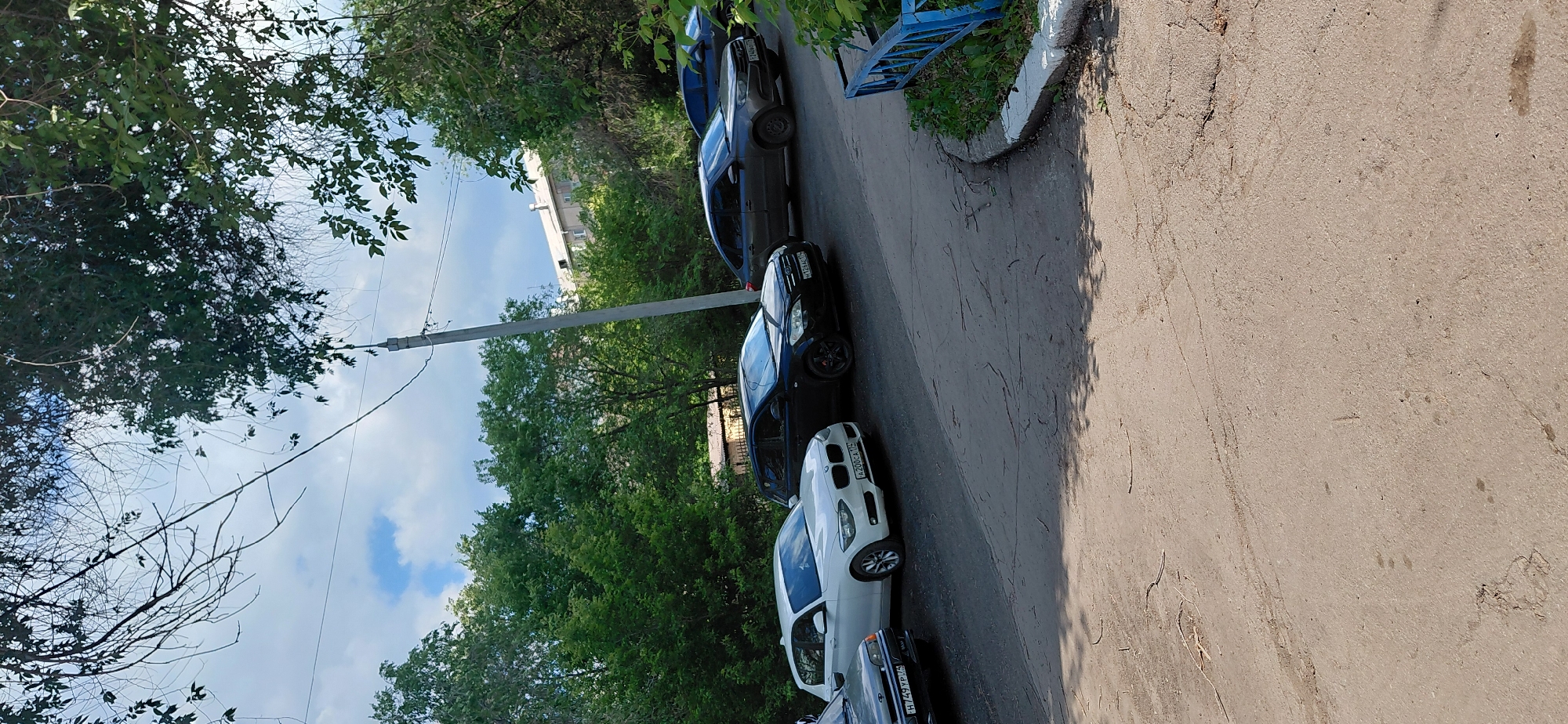 The image depicts an outdoor scene in Magnitogorsk, Russia, featuring a street lined with parked cars and dense green foliage. A light gray sedan is parked on the left, followed by a white hatchback, a black hatchback, and a dark gray sedan. The vehicles are parked on a paved surface that appears to be a road or parking area. Overhead, power lines are visible, strung between utility poles. The background is dominated by lush green trees, with a building visible in the distance. The sky is partly cloudy, with patches of blue sky and white clouds. A blue metal fence is present on the right side of the image, bordering a patch of grass and vegetation. The scene appears to be a residential or semi-residential area within the city.