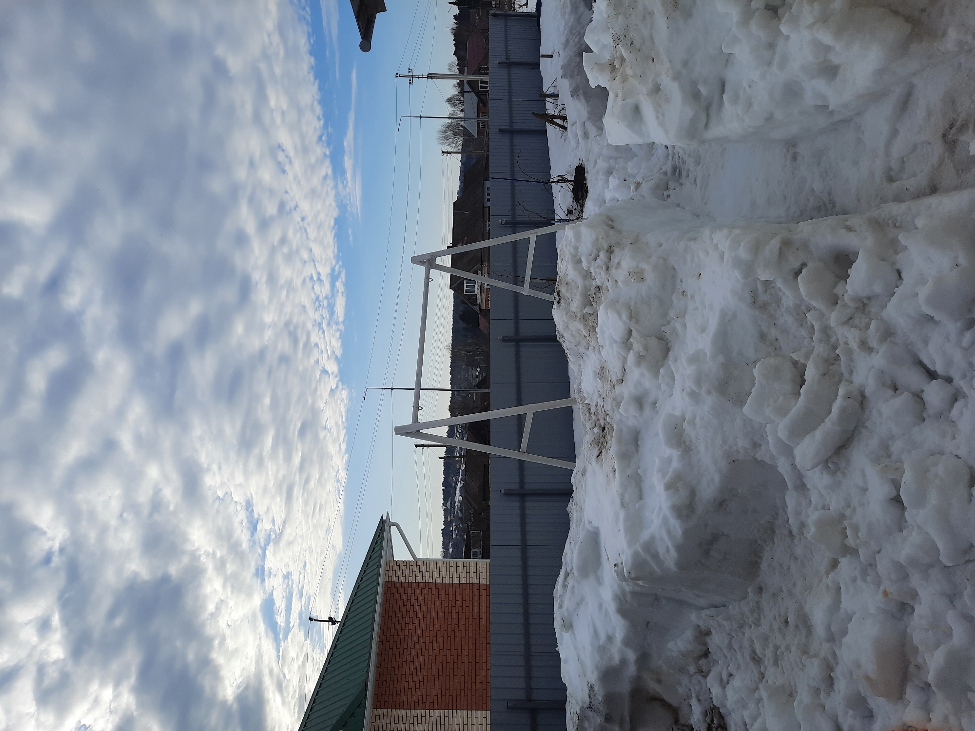 The image captures a winter scene in Krasnogorskoye, Russia. A gray fence stretches horizontally across the frame, separating a snow-covered foreground from a residential area in the background. Piles of snow, appearing blocky and textured, dominate the right side of the image. To the left of the fence, a portion of a brick building with a green roof is visible. A white metal structure stands near the fence, possibly a swing set frame. Above the fence, utility poles and wires traverse the skyline, and a few houses and trees are discernible in the distance. The sky is a mix of blue and clouds, creating a dynamic backdrop. The overall impression is one of a typical winter landscape in a suburban or rural setting.