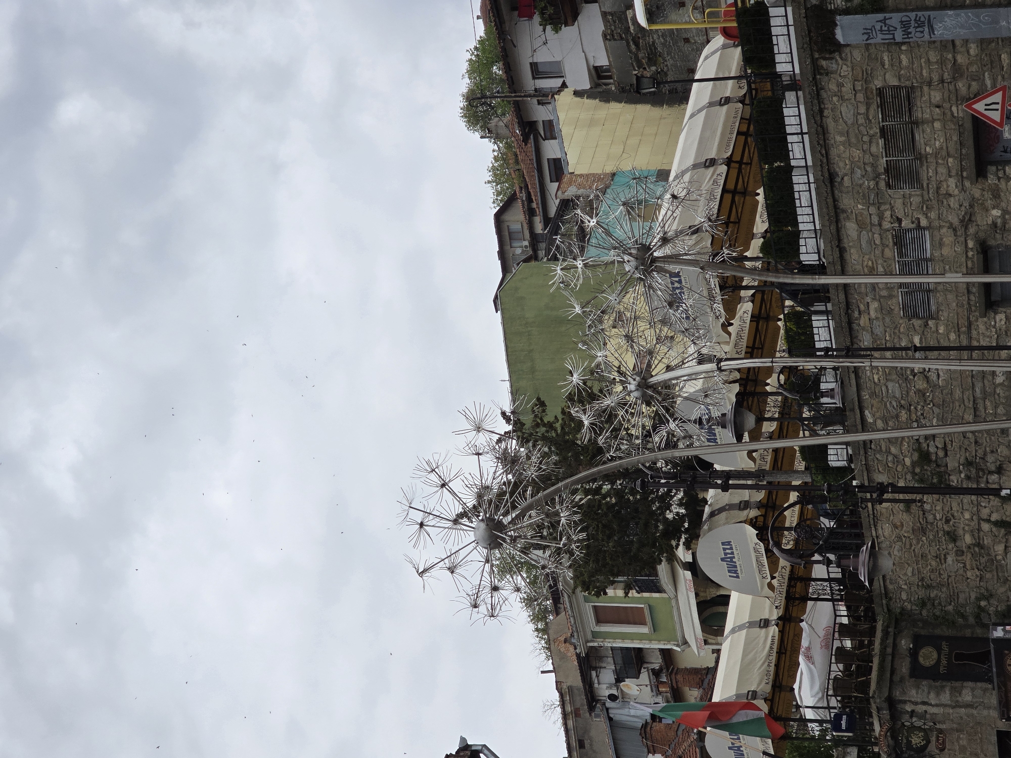 An outdoor urban scene depicts large, intricate metallic dandelion sculptures under an overcast, cloudy sky with several small birds flying. In the background, a cluster of buildings with varying architectural styles is visible. To the right, a multi-story building features a stone facade with rectangular windows, while other structures display painted exteriors in hues of green, yellow, and light blue.
An outdoor cafe or restaurant setup, frequently identified as "STRATILAT" (in both Latin and Cyrillic script: "СТРАТИЛАТ"), occupies the midground. It features numerous cream-colored awnings and umbrellas. Text visible on these awnings includes "LAVAZZA TORINO, ITALIA, 1995", "Coca-Cola", "КАФЕ-РЕСТОРАНТ" (Coffee-Restaurant), and "COFFEE RESTAURANT". Tables and chairs are arranged beneath the awnings, suggesting a public dining area, though no people are explicitly present.
Notable details include graffiti on a stone wall, which appears to read "LIAR HARD CORE" among other illegible markings, and a red triangular road sign indicating two-way traffic or a narrowing road. The overall weather is cloudy, suggesting daytime.