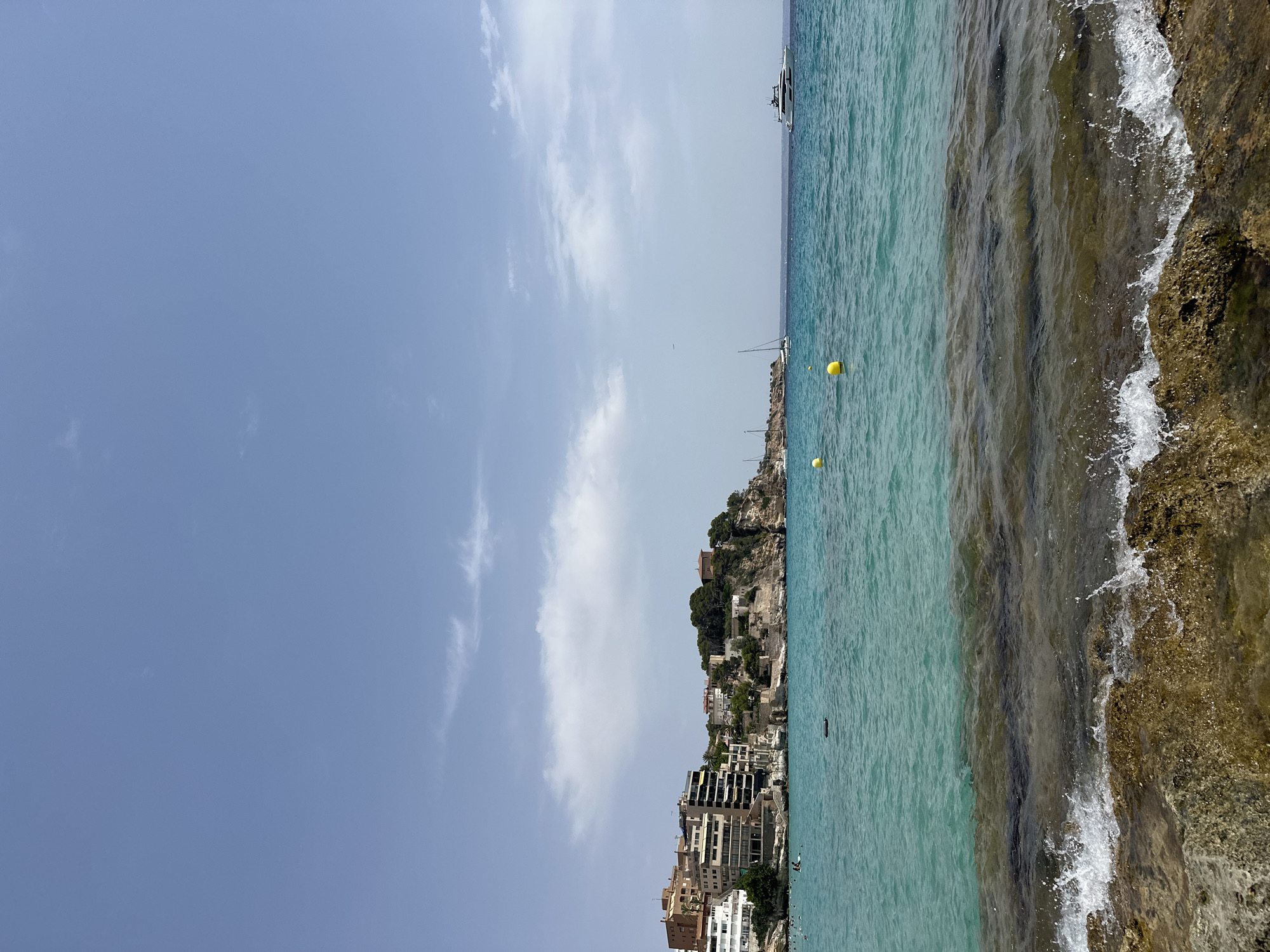 The image depicts a coastal scene in Sant Agustí, Spain, with a clear blue sky and scattered clouds. The foreground is dominated by rocky shore with waves crashing against it. The turquoise water extends towards the horizon, where several boats, including a large white yacht and sailboats, are visible. In the middle ground, a town with buildings nestled on a hillside is situated along the coast, characterized by a cluster of apartments and houses. A few yellow buoys are floating in the water. Scattered swimmers are present in the water near the shore, with one individual on a surfboard. The overall environment suggests a sunny day at a seaside resort.