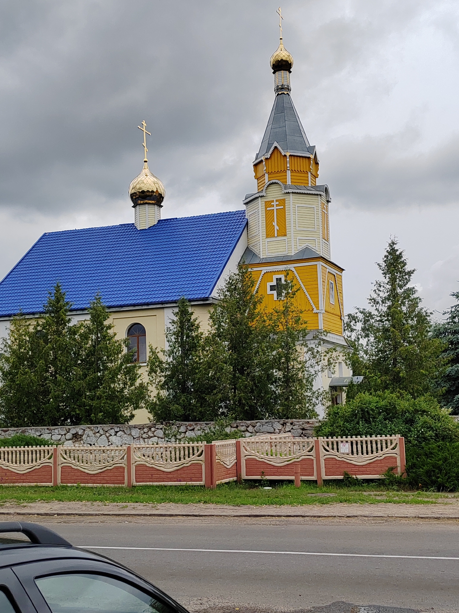 This is a medium shot of an Eastern Orthodox church in the village of Volchynsky Selsoviet, Belarus. The church has a blue metal roof and a tall, yellow bell tower with a gray spire. The bell tower is topped with a golden dome and cross. The church is surrounded by trees and a decorative fence. The sky is cloudy. A portion of a dark-colored car is visible in the foreground, obscuring a segment of the road and sidewalk.
