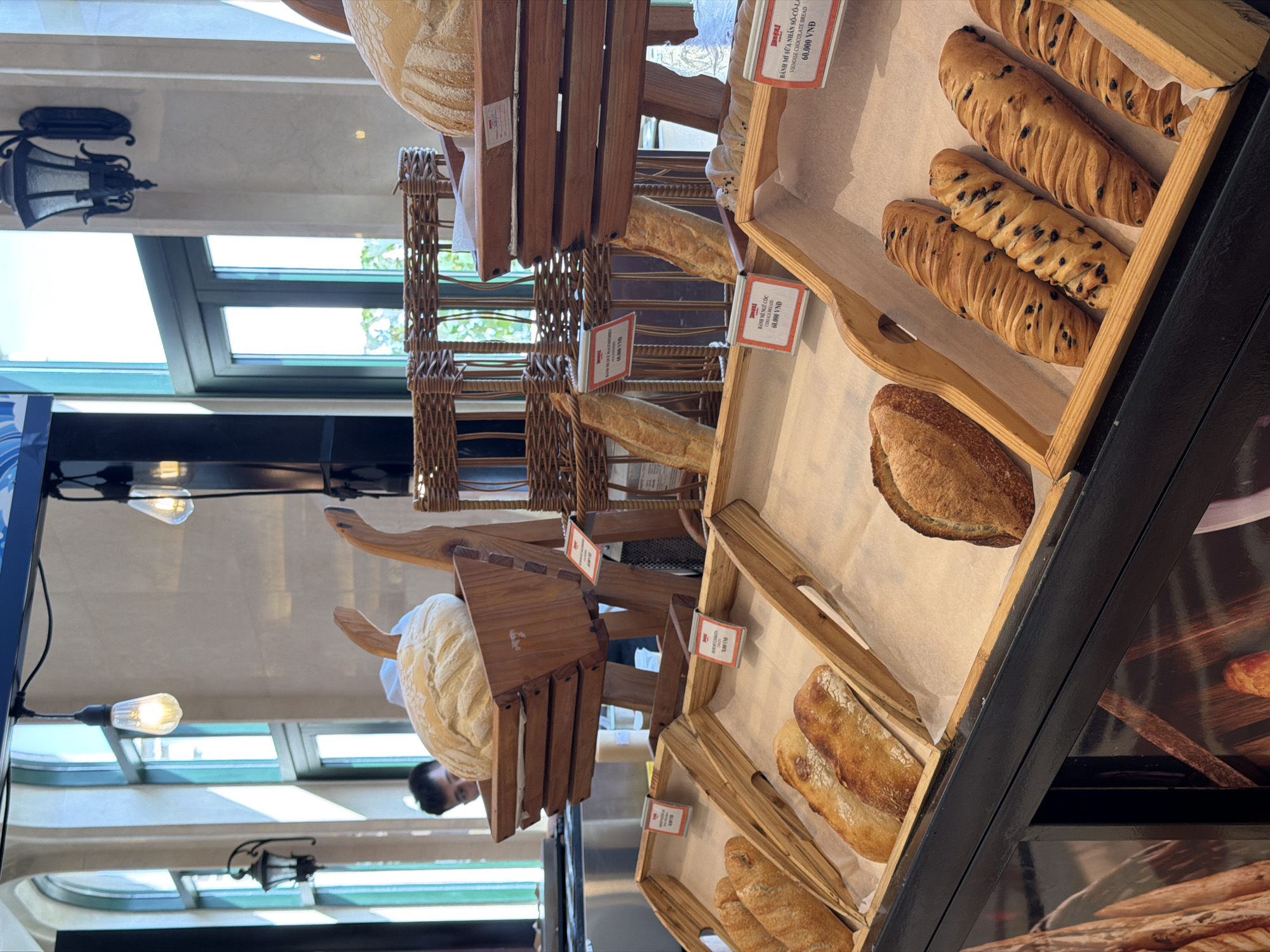 A multi-tiered wooden display case filled with various baked goods, including baguettes, loaves of bread, and pastries with chocolate chips, dominates the scene. Price tags, displaying "60,000 VND" for each item, are visible. The camera angle gives a view of the breads from above in a bakery in Da Nang, Vietnam. A person is visible in the background. The bakery has large windows and decorative lighting.
