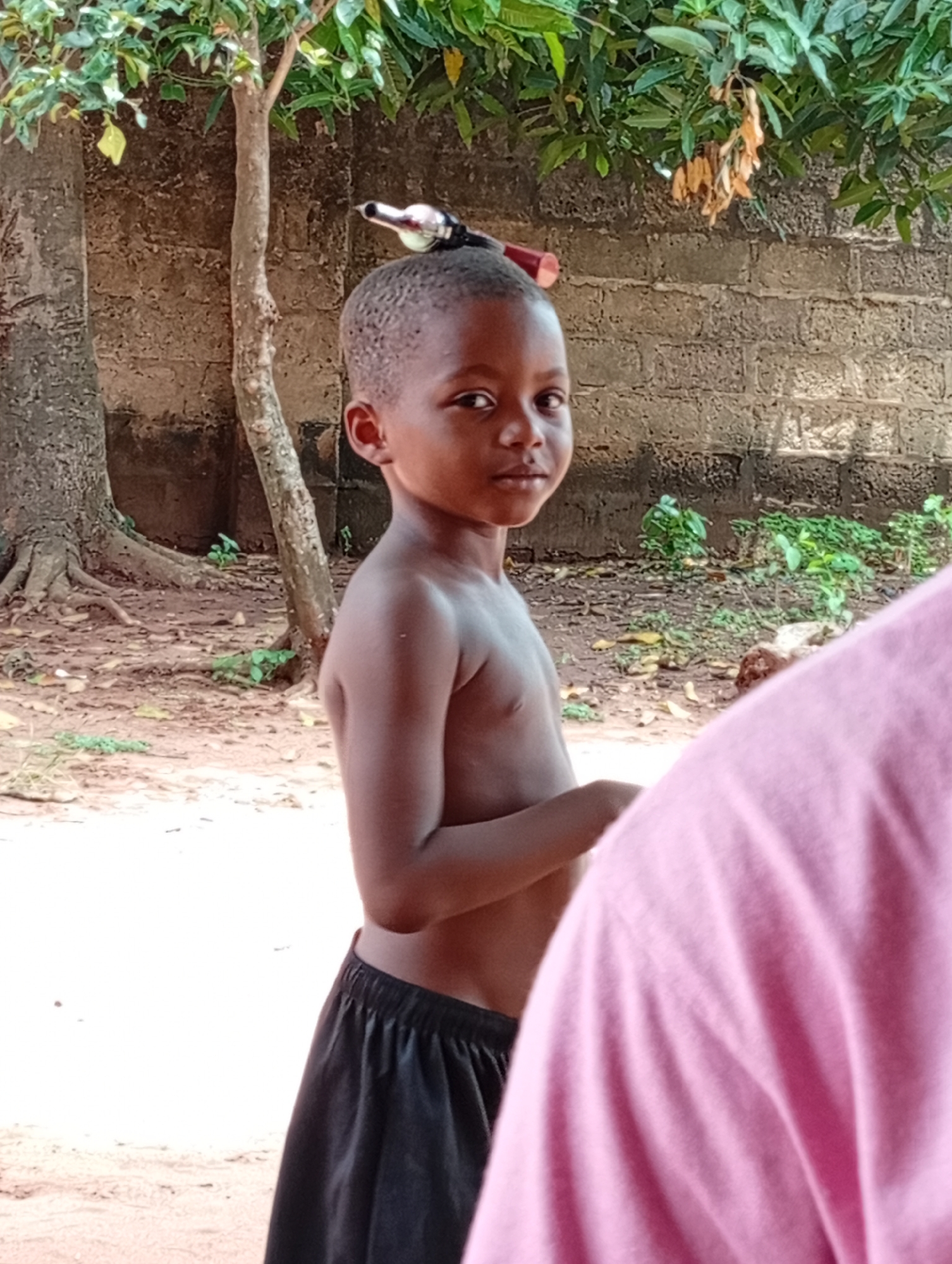 A young, bare-chested boy stands in what appears to be an outdoor area in Ogidi, Nigeria. He has short hair and is wearing black shorts. A small object, possibly a bottle opener, is balanced on his head. Behind him is a tree and a brick wall, while in the foreground, a pink-shirted person is partially visible. The ground is dry and dusty.