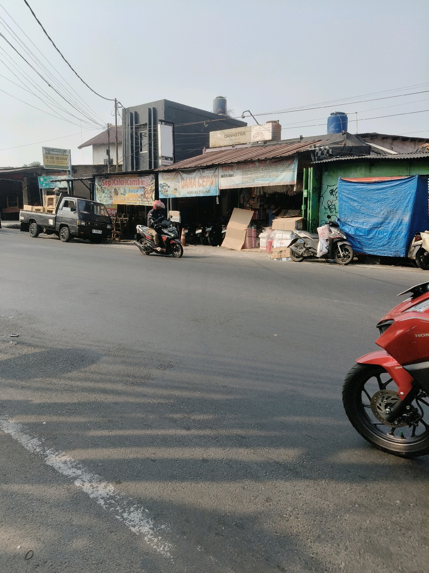 The image depicts a street scene in South Tangerang, Indonesia, likely in a commercial or semi-industrial area. A grey Mitsubishi pickup truck is parked on the left side of the road, loaded with several boxes. To the right of the truck, a person is riding a red and white motorcycle, wearing a helmet and casual clothing. Further down the street, a row of low-rise buildings lines the road, housing various businesses. Signs visible include "SOP KAKI KAMBING," "DANA CEPAT," and "JUANAYA PLASTIK." The buildings have corrugated metal roofs, some with water tanks. A large blue tarp is draped over a section of one building. In the foreground, the front wheel and fender of a red motorcycle are visible. The road surface is asphalt, with a broken white line marking the lane. The sky is clear and pale blue, suggesting daytime. Overhead, a complex network of electrical wires crisscrosses the scene.