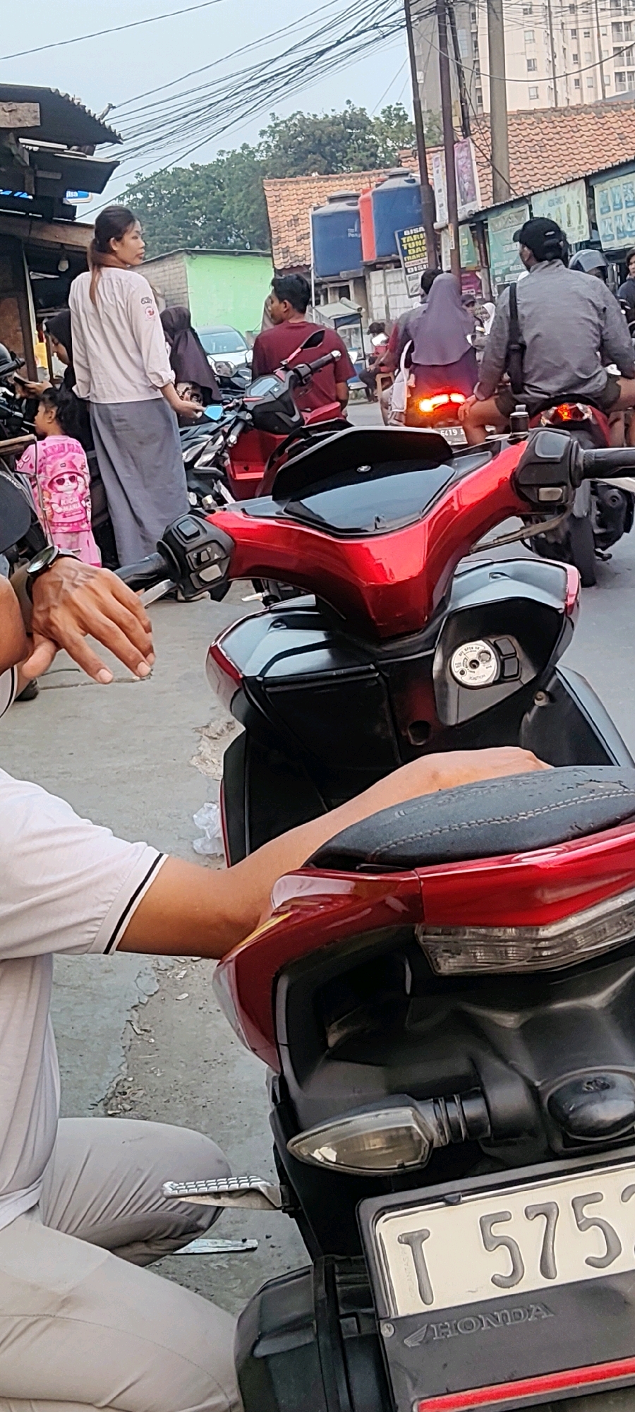 The image depicts a street scene in Mekarmukti, Indonesia, characterized by its urban environment and the presence of motorcycles. In the foreground, a red and black motorcycle is prominently featured. The handlebars and front fairing of the motorcycle are visible, with a key ignition switch on the panel. A license plate reading "T 5752" with "HONDA" below it is attached to the rear of a similar motorcycle in the foreground.
Several people are present in the scene. A person wearing a white shirt with black trim and grey pants is seated in the lower left portion of the image, with their arm resting on the motorcycle seat. Further back, a woman with long dark hair tied in a ponytail, wearing a light-colored long-sleeved shirt and grey pants, stands and looks to the left. A young child in a pink outfit with cartoon characters is also visible near her. Several other individuals are seen on or near motorcycles in the background, some wearing helmets and headscarves.
The environment appears to be a street with overhead wires and utility poles. Buildings with tiled roofs and some with exposed brick are visible in the middle ground, along with large blue water tanks. In the far background, a tall building suggests an urban setting. The overall atmosphere suggests a bustling street, likely during daylight hours given the lighting conditions.