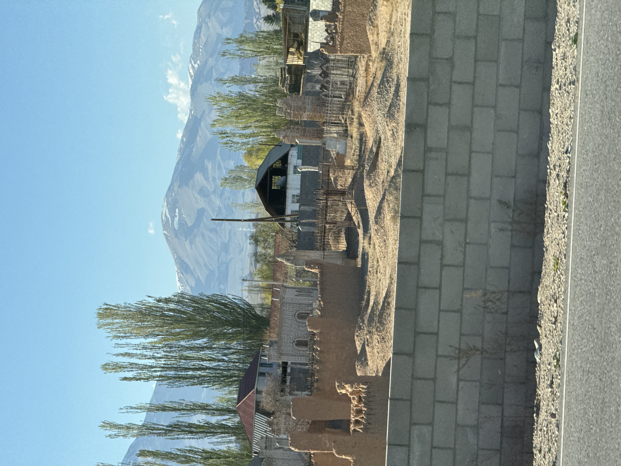 The image shows a view in Toru-Aigyr, Kyrgyzstan. A mountain rises in the background, partially obscured by trees. In the foreground are structures, some in disrepair, including buildings with metal roofs and brick or stone walls. A concrete block wall runs along the bottom right, next to a paved road or pathway. The sky is blue with scattered clouds. The overall setting appears to be a rural or semi-rural area.