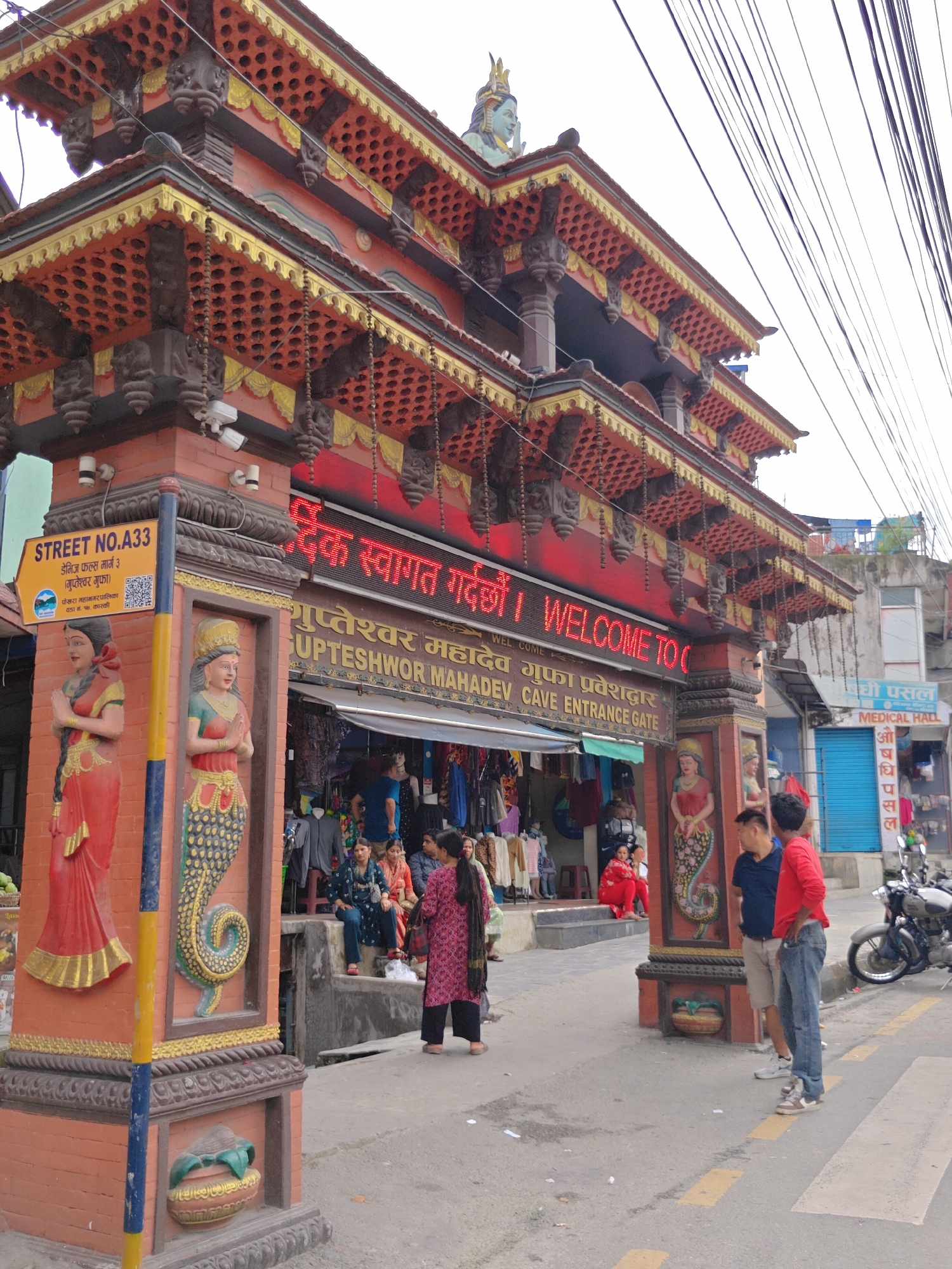 A wide-angle, eye-level shot captures the ornate entrance gate to Gupteshwor Mahadev Cave in Pokhara, Nepal. The gate, constructed primarily of brick and adorned with intricate wood carvings and gold accents, features multiple tiered roofs. A prominent red LED sign above the entrance reads, in both Nepali and English, "WELCOME TO GUPTESHWOR MAHADEV CAVE ENTRANCE GATE." Flanking the entryway are two statues of women in traditional attire, one on each side.
In the foreground, to the right, two young men stand on the street, one in a blue t-shirt and khaki shorts, the other in a red long-sleeved shirt and jeans. They appear to be conversing. A motorcycle is parked on the right side of the street. A crosswalk with yellow stripes is visible on the asphalt road.
Behind the gate, a bustling marketplace is visible, with various stalls displaying clothing and other goods. Several people, mostly women, are seen browsing or sitting near the shops. The sky above is overcast. To the far right, a sign for "MEDICAL HALL" is visible. The overall scene depicts a lively streetscape at a significant cultural landmark in Pokhara.