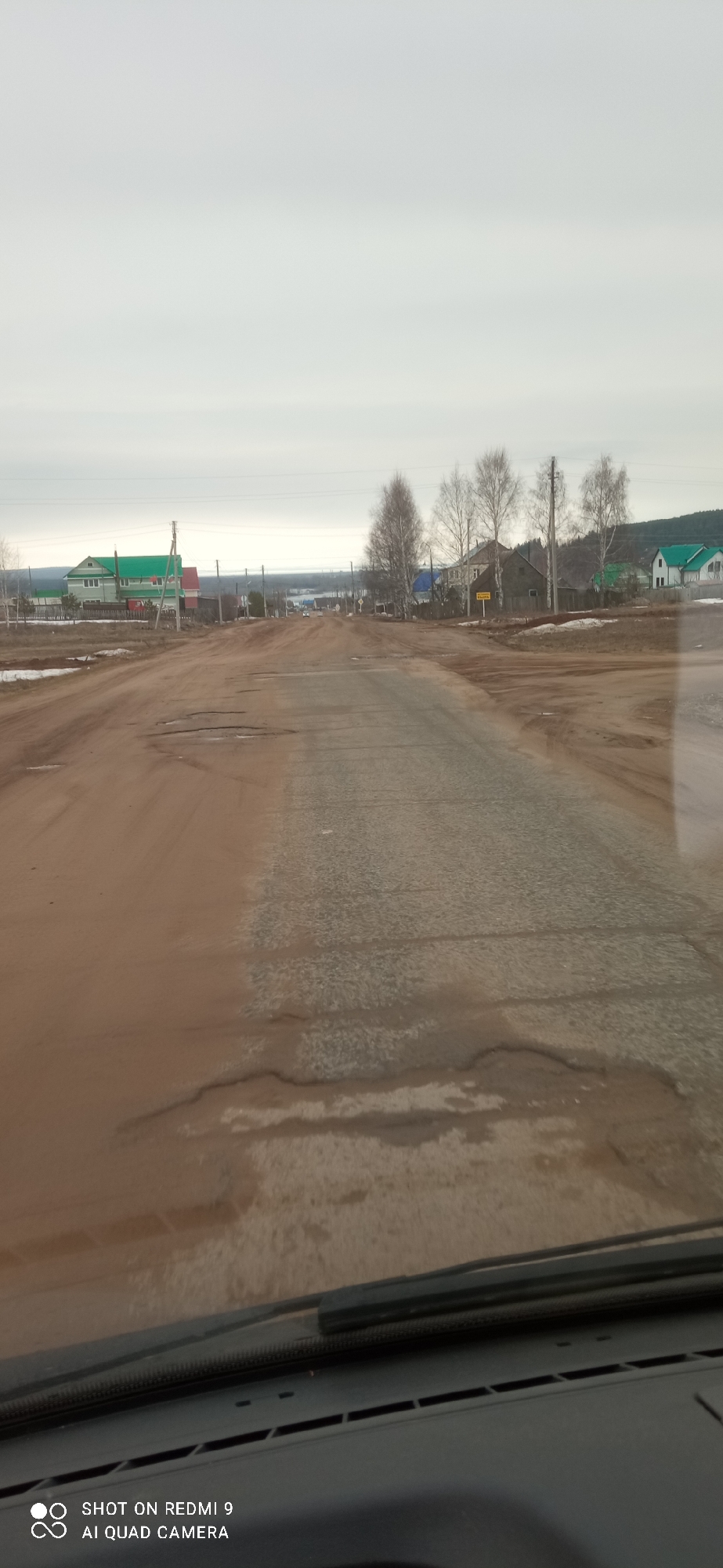 The image depicts a rural street in Ольховка, Russia. The view is from inside a vehicle, likely a car, looking out through the windshield. The road is predominantly dirt, with sections of worn asphalt visible. Puddles and potholes are scattered across the road surface. Buildings, likely houses, are visible on either side of the road. Some have green roofs, and there are bare trees interspersed amongst them. Power lines are present, running alongside the road. The sky is overcast, suggesting a dull or gray day. The dashboard of the vehicle is visible at the bottom of the image.