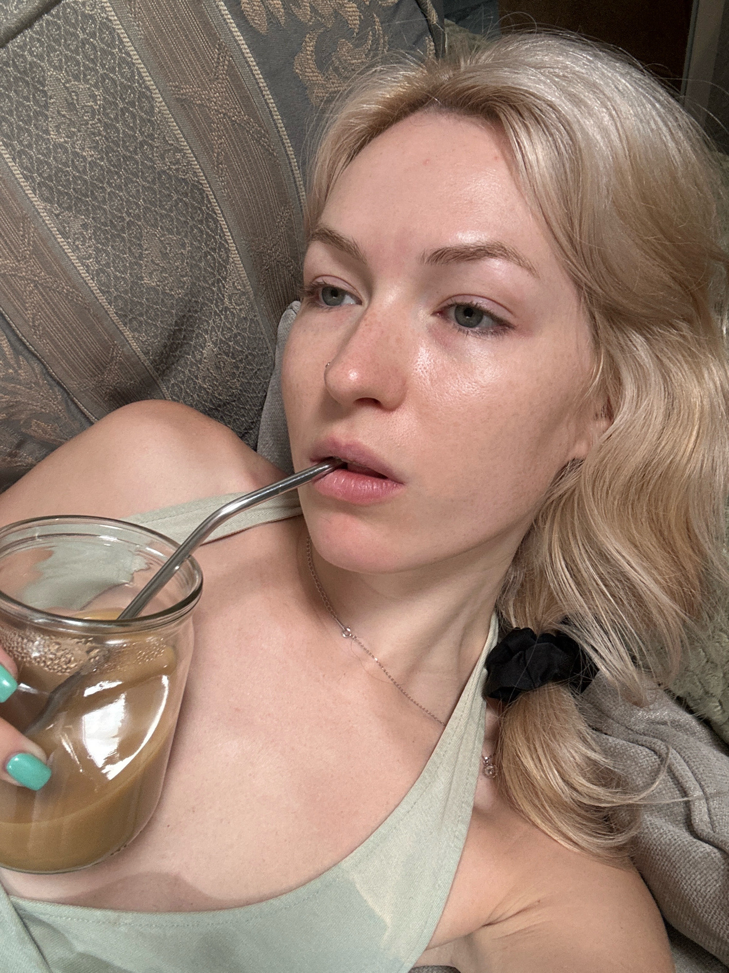 This is a close-up, low-angle selfie of a young woman with blonde hair and light skin, lying down and drinking from a glass jar with a metal straw. She is wearing a light green tank top and a silver necklace. Her fingernails are painted a bright turquoise color. The background appears to be a textured sofa with a patterned fabric. The scene is indoors. The image does not provide any information about the city of Tbilisi, Georgia.