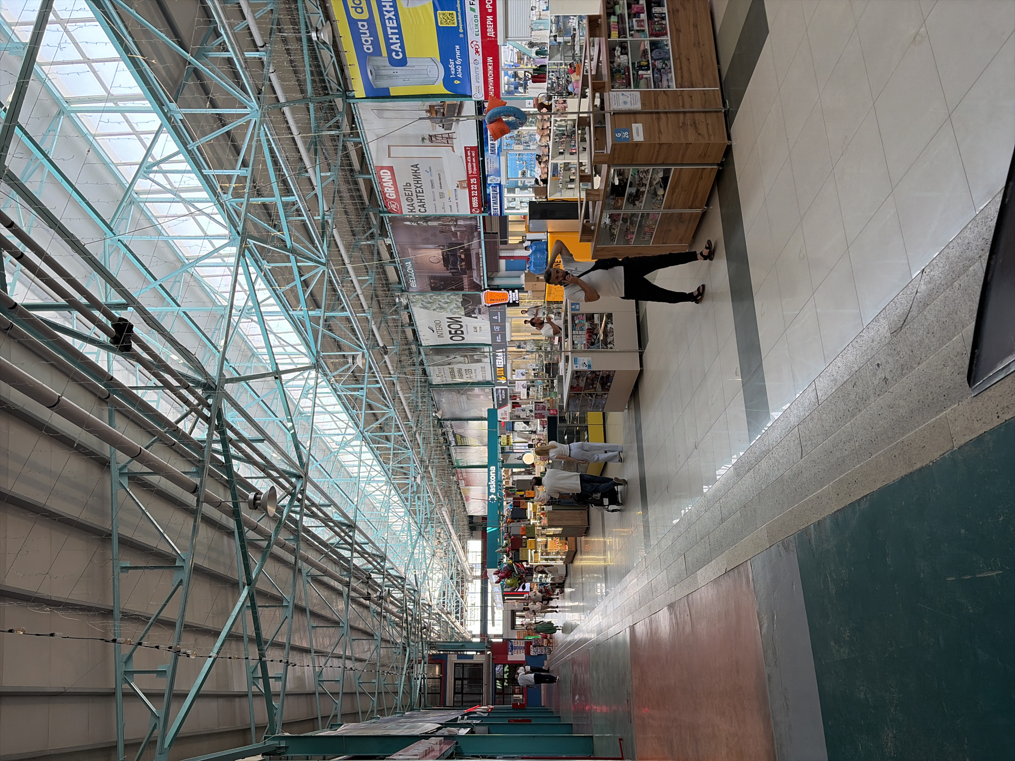 The image shows an interior view of a large, modern marketplace, likely a shopping mall or bazaar, in Bishkek, Kyrgyzstan. The ceiling is constructed with a metal framework and translucent panels that allow natural light to filter in. Various stalls and shops are visible along the wide, tiled walkway, displaying a range of goods. Some stalls have glass display cases, while others showcase items on shelves. Prominent signage in Russian indicates brands like "ASKONA" and advertisements for home goods and plumbing supplies. Several people are present in the scene: a man in a white shirt and black pants is walking away from the camera, another man in a white t-shirt and black pants is standing near a stall with his hand on his head, and a couple with a stroller is walking down the aisle. Other individuals are seen browsing or working in the background. The overall impression is a busy, well-lit commercial environment.