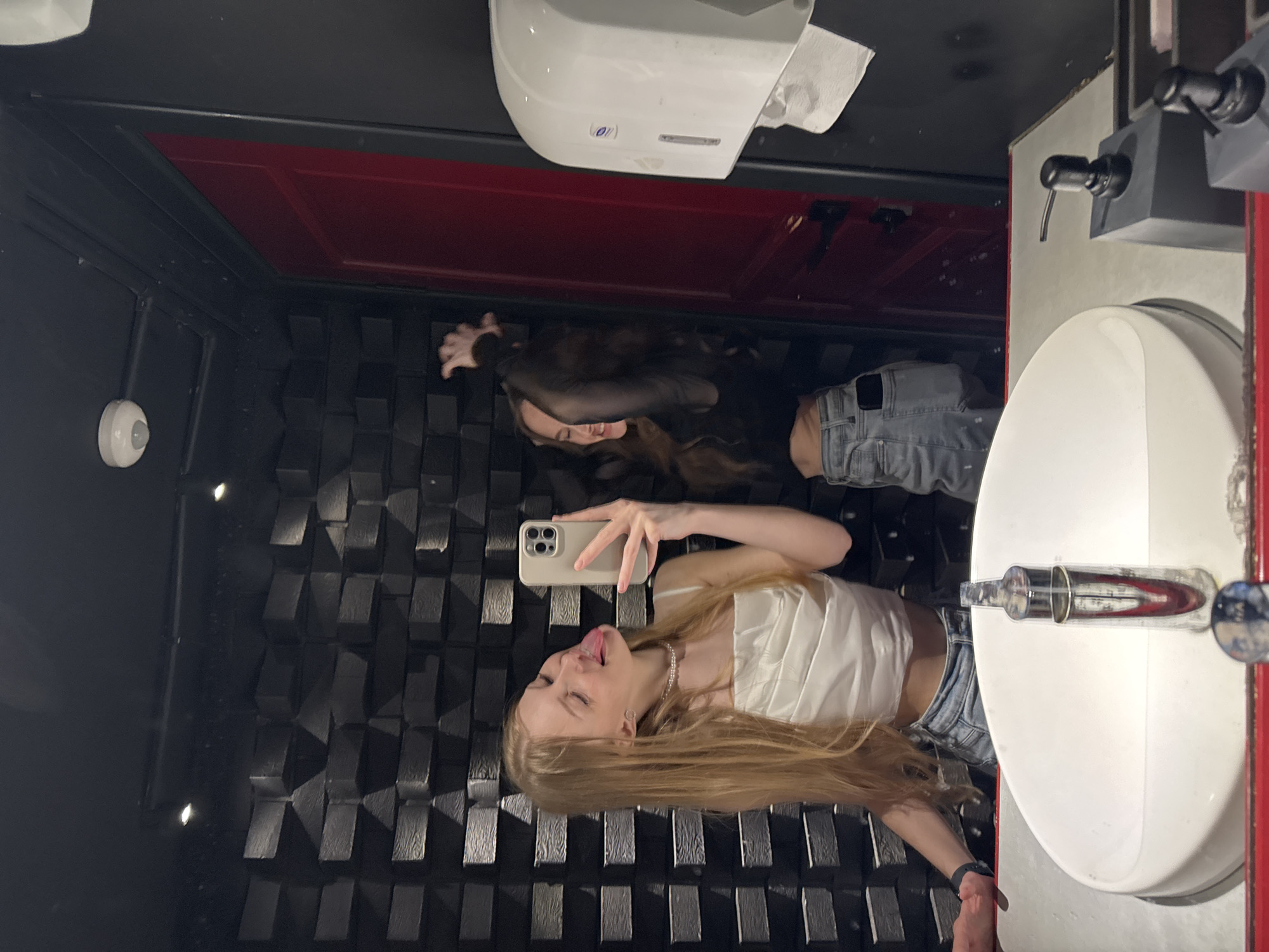 The media file is a vertical, mirrored, low-angle, indoor photograph, likely taken in a restroom in Perm, Russia. Two young women are the primary subjects, reflected in a large mirror that covers the entire background. The setting appears to be a modern, darkly-themed restroom with textured black walls.
In the foreground, a blonde woman is leaning forward, sticking her tongue out playfully as she holds up a smartphone to take a selfie. She is wearing a white ruched-top and denim shorts, accessorized with a pearl necklace and a black wristband. Her hair is long and blonde, cascading around her.
Behind her, another woman with long, dark hair is visible in the reflection. She is wearing a black sheer top and denim jeans. Her expression is a smile.
Objects in the scene include a white paper towel dispenser mounted on the wall above the dark textured surface, a sink with a chrome faucet, and a red door. The lighting is dim, suggesting it might be nighttime or the restroom has mood lighting. The overall mood is playful and lighthearted, indicated by the facial expression and pose of the blonde woman. No visible text is present in the image.