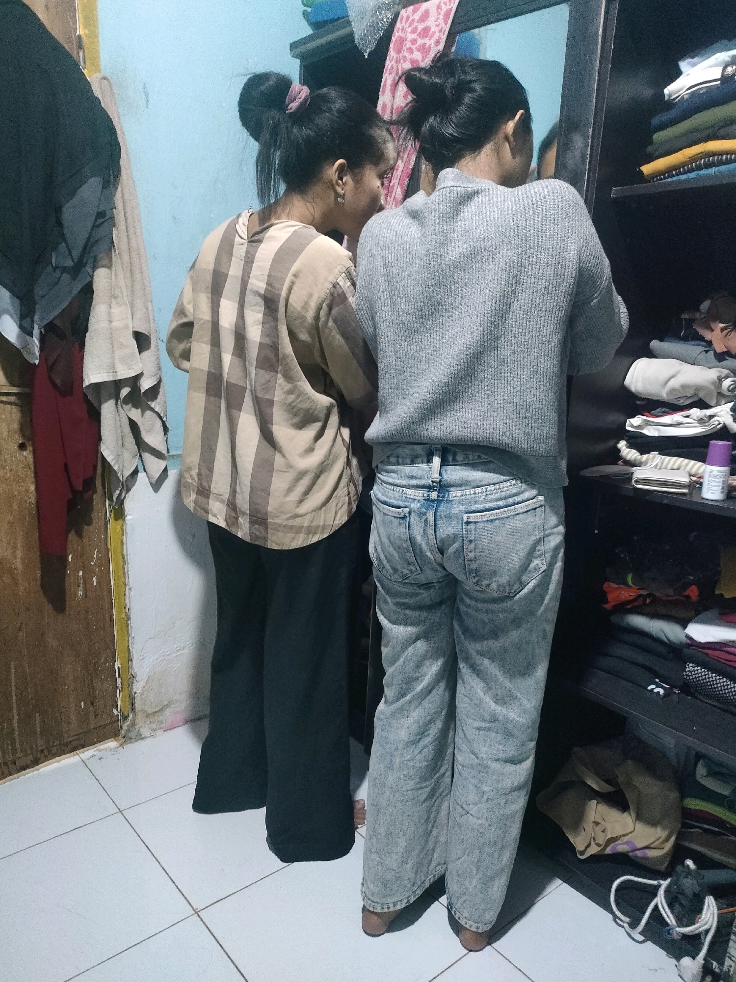 In a room with a tiled floor and light blue walls, two young women are standing. The woman on the left wears a beige and brown plaid shirt and black pants. The woman on the right wears a grey sweater and light blue acid-wash jeans. They are both facing away from the camera, towards a black wardrobe with shelves filled with folded clothes. The wardrobe is open, and a mirror is attached to its door, reflecting the room. On the floor next to the wardrobe, there is a beige bag and a white rope. To the left of the women, a wooden door is visible, with some clothing hanging on it and on the wall next to it. The scene appears to be a bedroom or a dressing area within a residential setting in Pekanbaru, Indonesia.