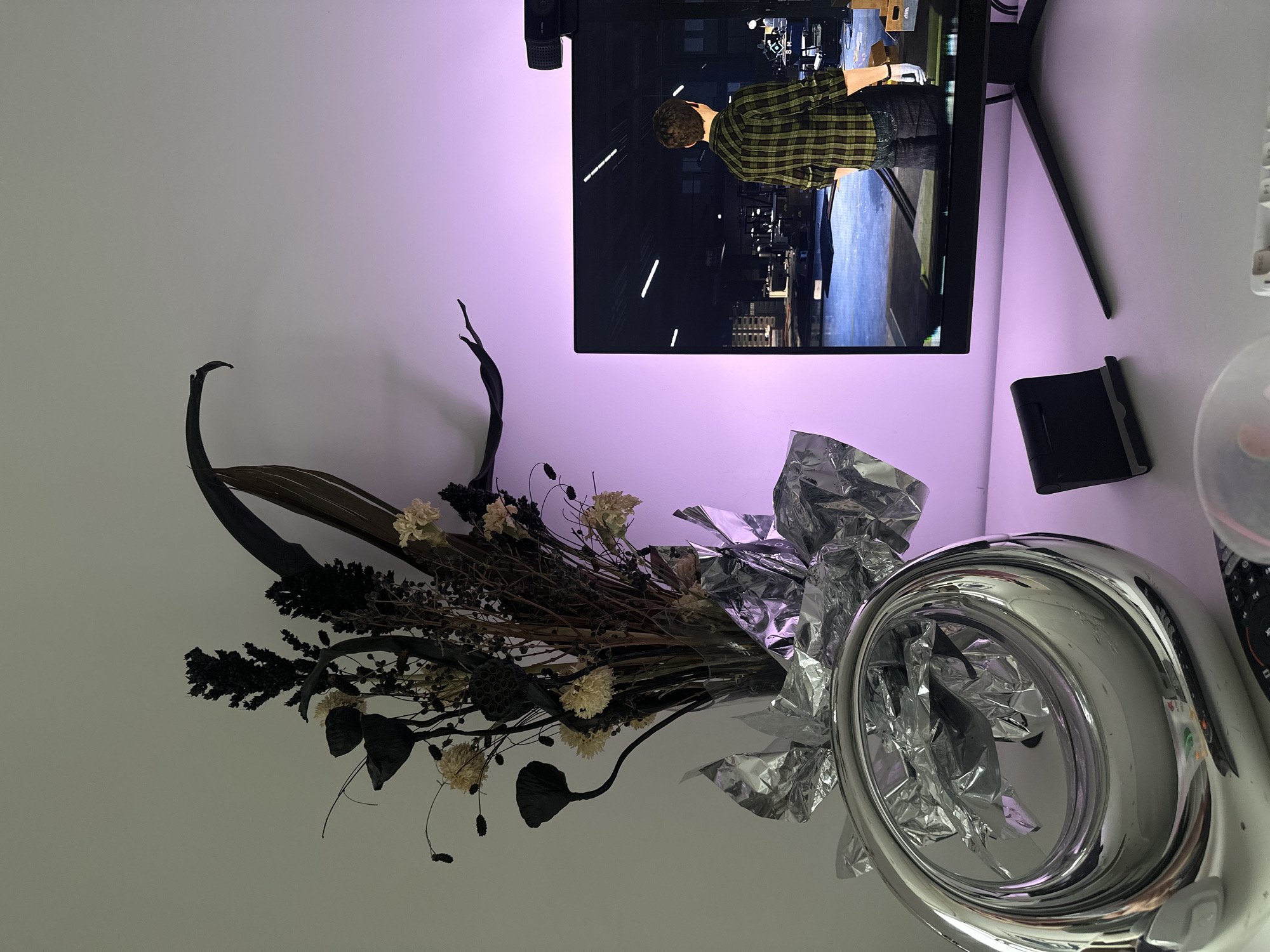 The image depicts a still life arrangement against a white wall illuminated with a purple hue. A black computer monitor is situated at the upper center of the frame, with a man in a green and yellow plaid shirt visible on the screen. A black webcam is affixed to the top of the monitor. To the right of the monitor is a small black electronic device. Below the monitor and to the left is a dried floral arrangement in a metallic silver vase. The arrangement includes dark, dried branches and flowers. The vase is partially filled with crumpled silver foil. The photo was likely taken indoors. There is no evidence to suggest the location is in Moscow, Russia.