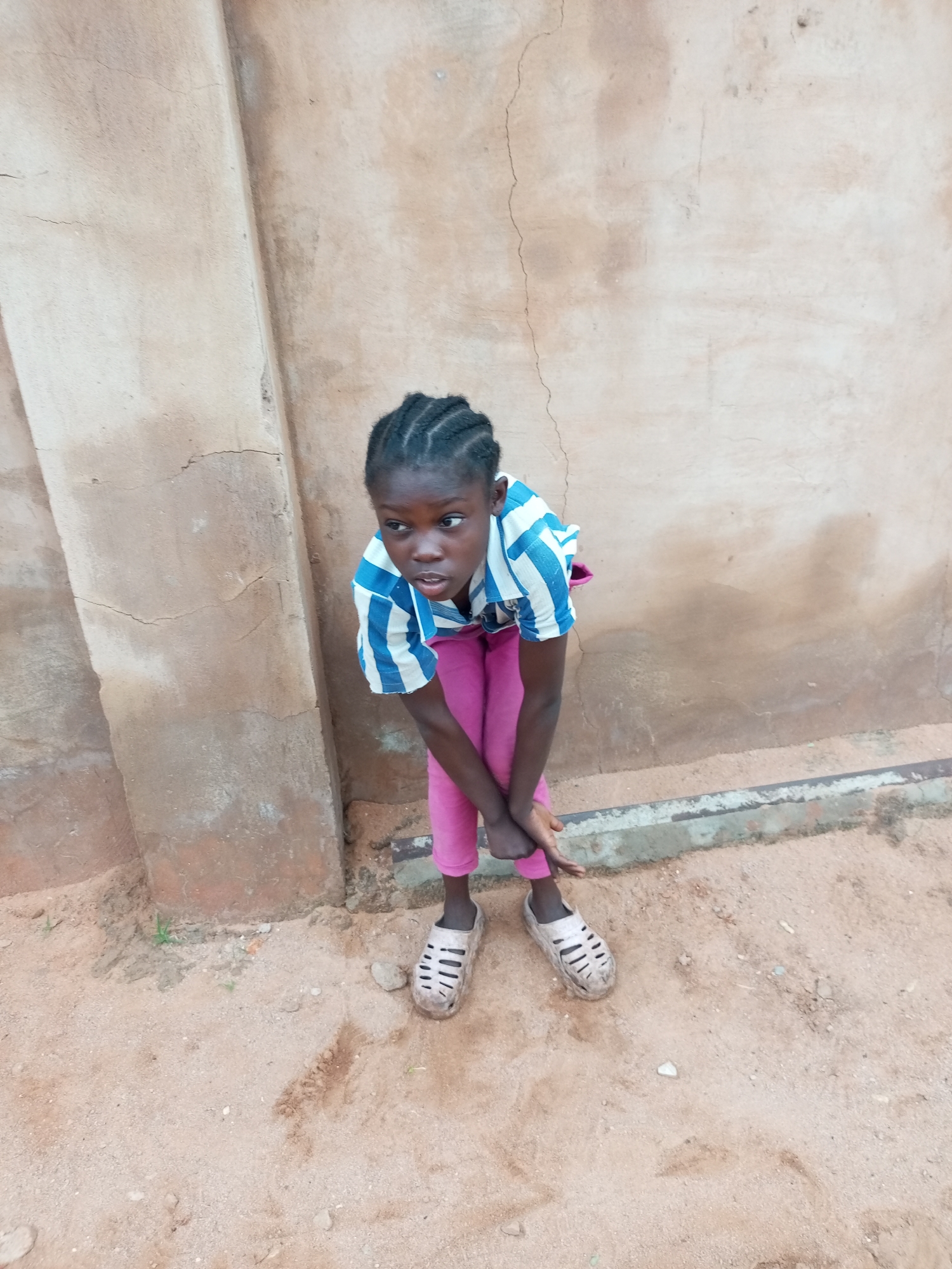 A young Black girl with braided hair is crouched down against a textured, light brown wall in Tempure, Nigeria. She is wearing a blue and white striped shirt and pink pants, along with dirty beige sandals. The ground is sandy with scattered pebbles. The wall shows signs of wear, including cracks and discoloration.