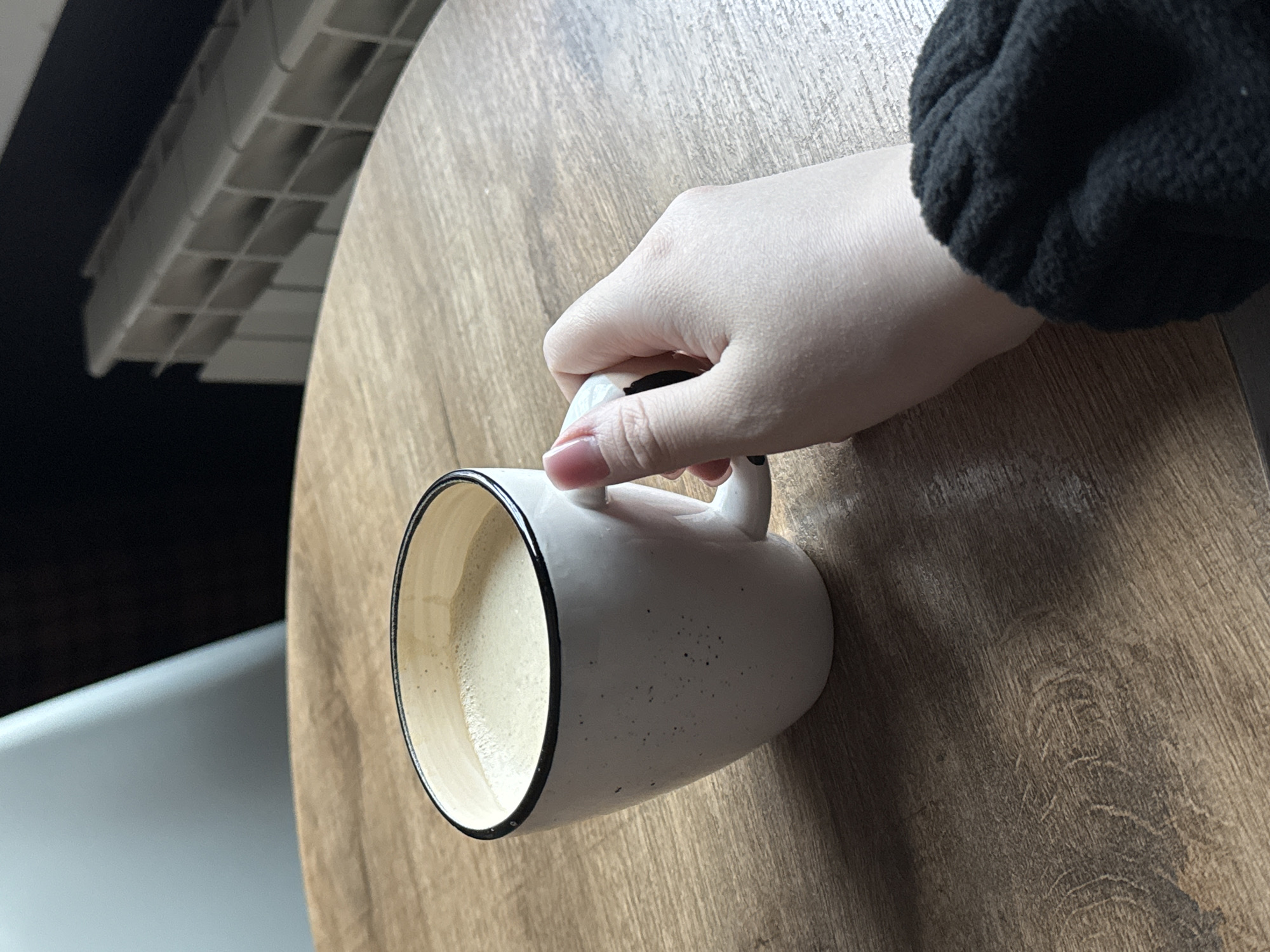 A close-up shot depicts a person's hand holding a white mug with a black rim, partially filled with a light-colored liquid, possibly coffee or tea. The mug sits on a wooden surface. The person's arm, clad in a black sweater, is visible in the upper right corner. To the left, a white object with a geometric pattern is seen, possibly a part of furniture or decor. The location appears to be indoors, possibly a cafe or a home in Unknown, Unknown.