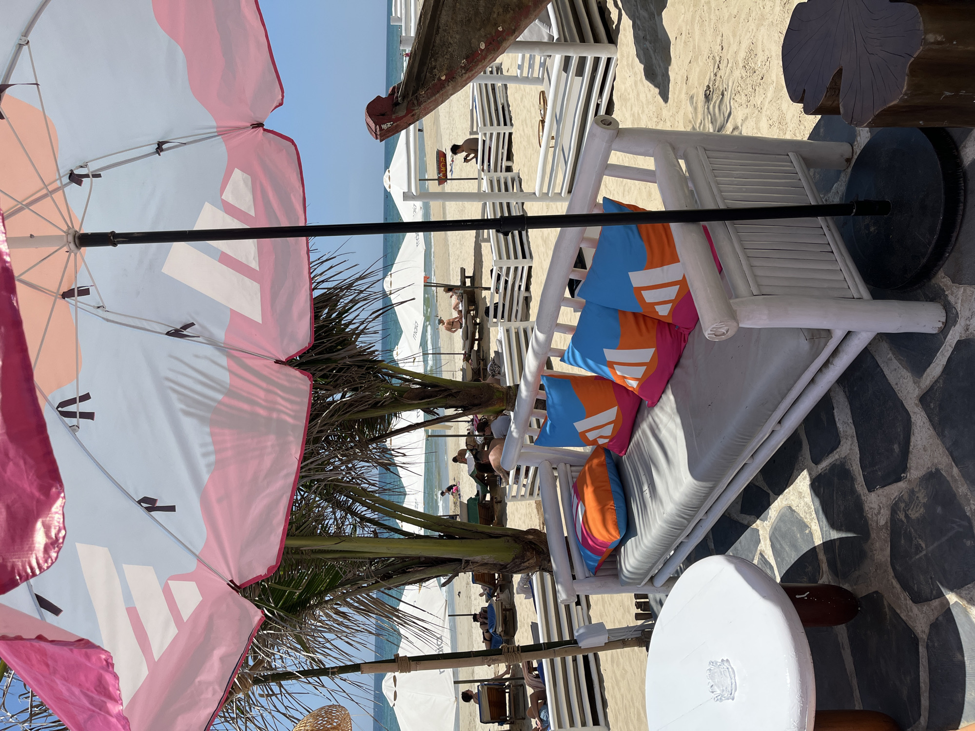 This is a photo of a beach scene, likely taken from an elevated perspective under a beach umbrella, possibly in Đà Nẵng, Vietnam.
The scene includes:
* A beach umbrella with a pink and white patterned top
* A white beach lounge with colorful pillows
* White beach chairs and tables
* Palm trees
* A beach with people, umbrellas, and the ocean in the background.
* There is a boat-shaped object in the background.
* There is a low white round table in the foreground.
* The ground is paved with irregular black stones.