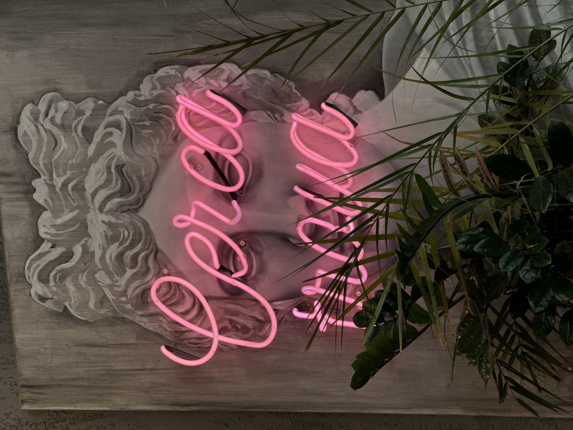 The image presents a close-up, slightly angled view of a painted bust of a classical figure. The bust, rendered in grayscale, is overlaid with a neon sign in cursive pink. The sign's script reads "Dream Team". The painting is on a textured surface, possibly a canvas or wall, with a gray-brown tone. Green plants, including leafy foliage and slender palm-like fronds, frame the right side and partially obscure the bust and signage. The scene is located in Perm, Russia.